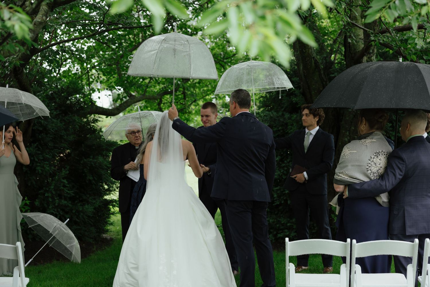 Outdoor rainy wedding ceremony with guests holding clear umbrellas as bride walks the aisle.