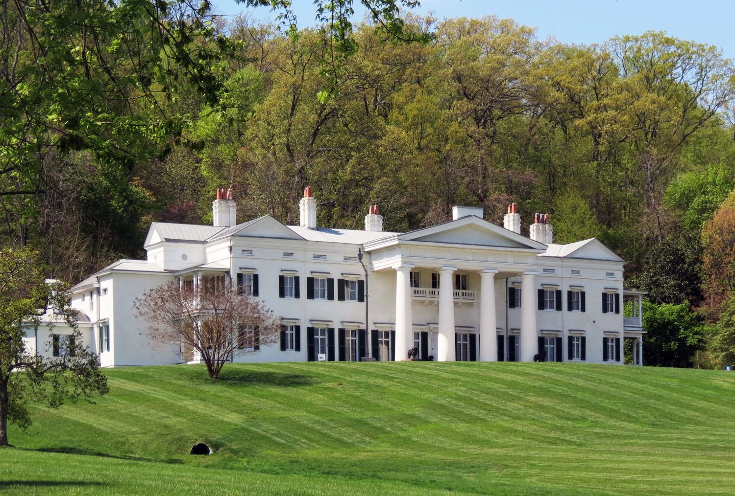 Large white neoclassical mansion with columns, black shutters, and manicured green lawn surrounded by lush spring trees.