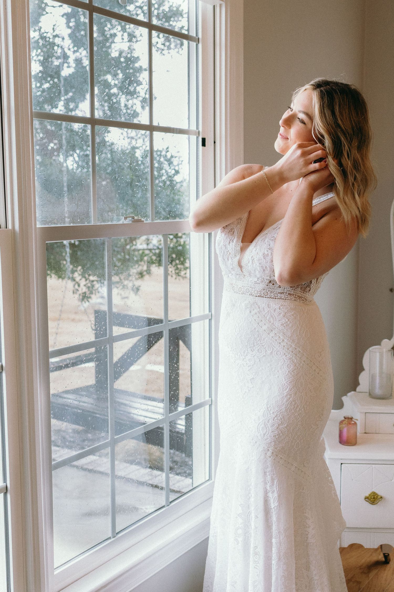 Bride in white lace wedding gown standing by a sunlit window, adjusting her necklace with trees visible outside.