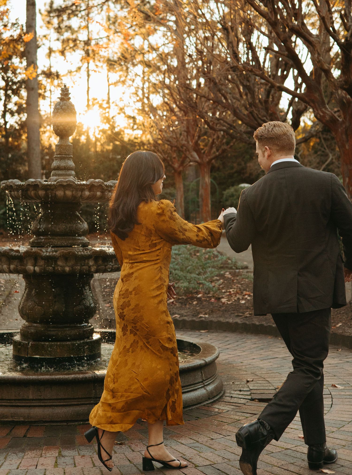 Woman in mustard dress and man in dark suit walking hand in hand toward a garden fountain at sunset.