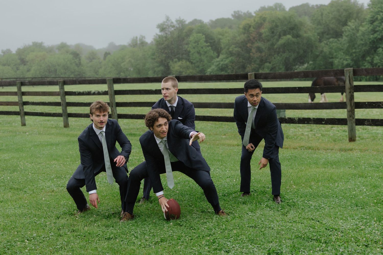 Four groomsmen in dark suits playfully posing with football on rainy green farm field.