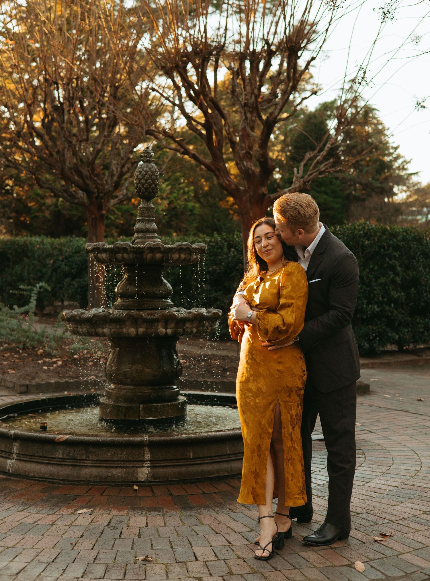Couple embracing tenderly by a tiered stone fountain in an autumn garden at golden hour.