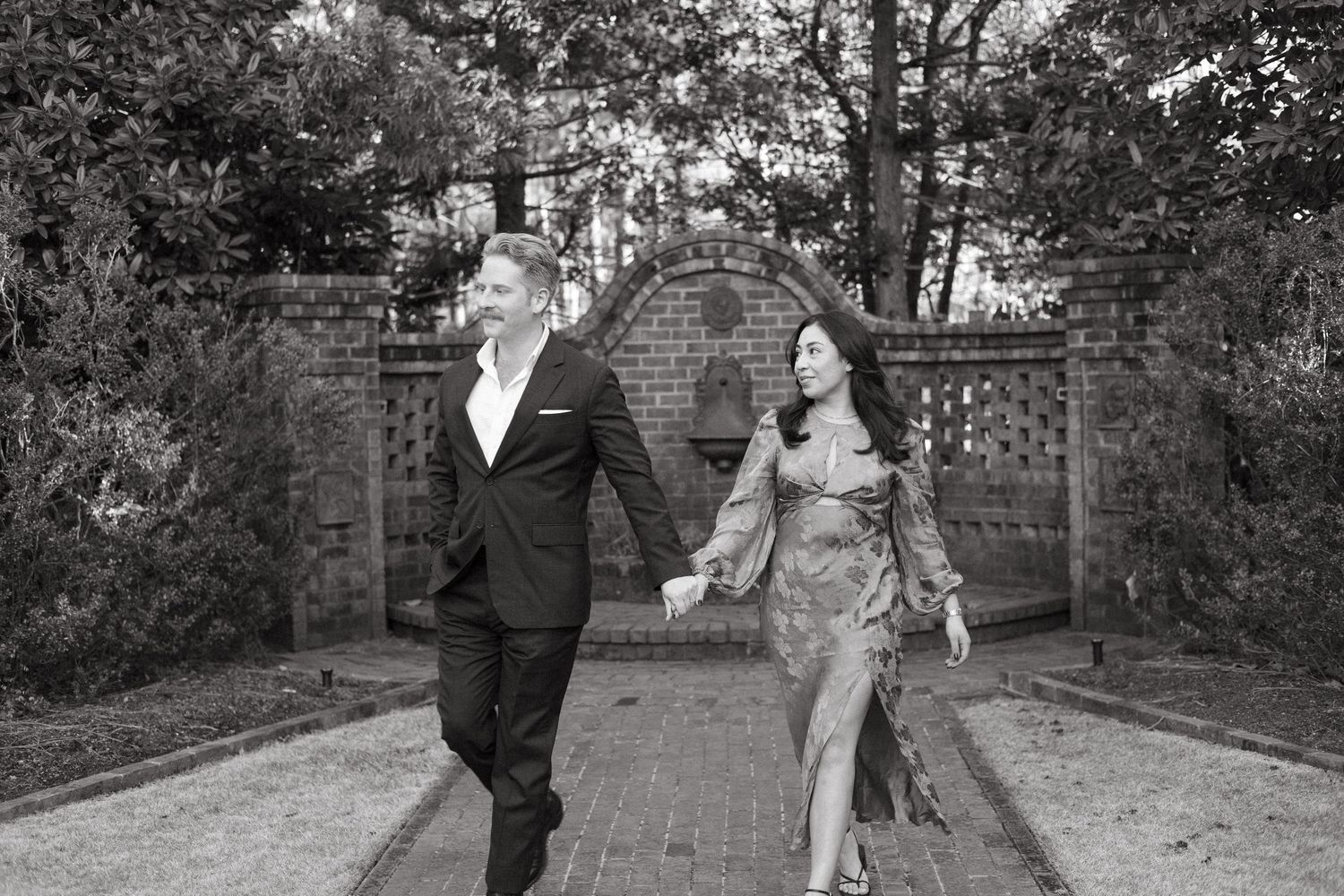 Black and white photo of couple walking hand in hand through brick garden pathway gate.