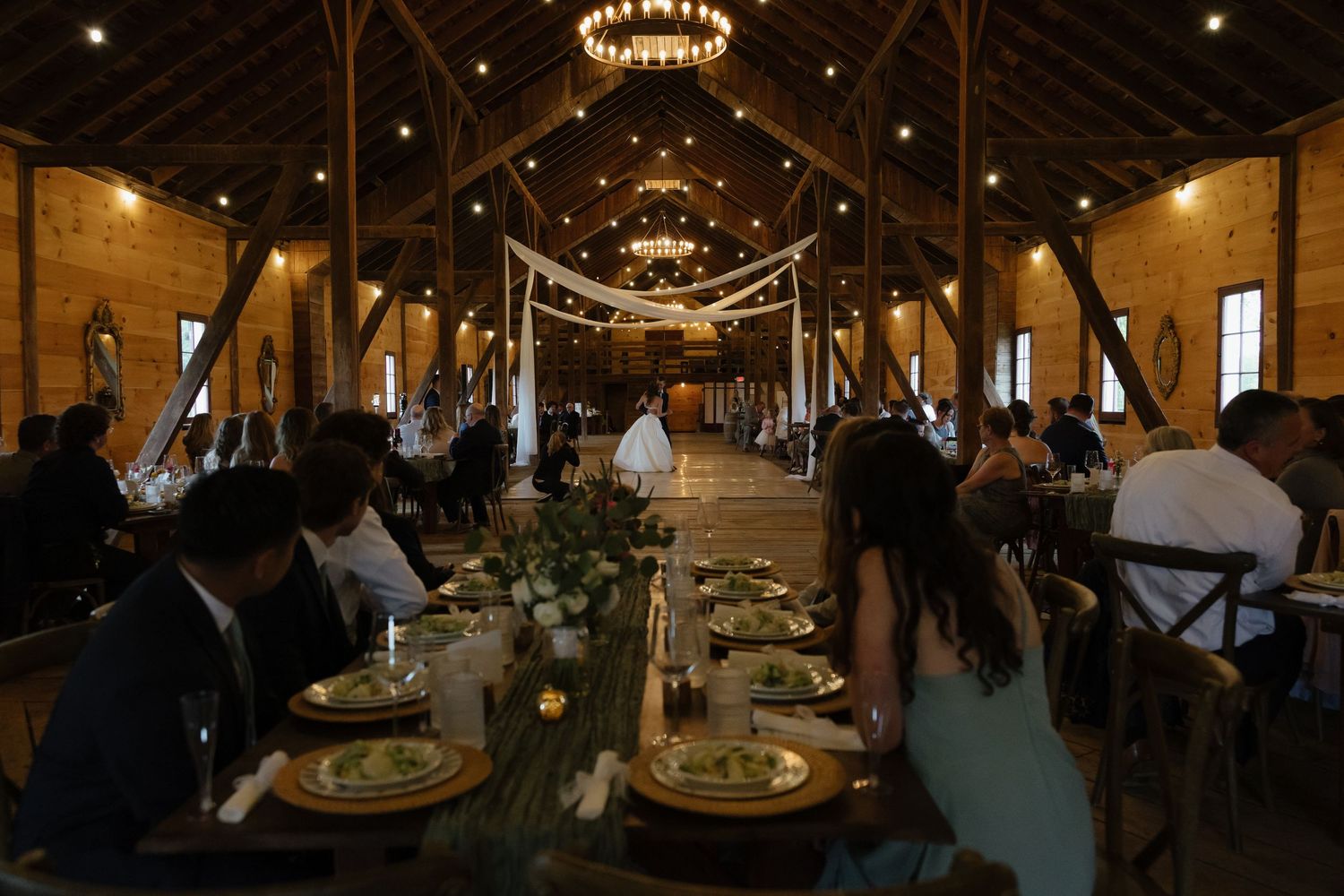 Elegant barn wedding reception with long wooden tables, greenery centerpieces, and warm chandelier lighting.