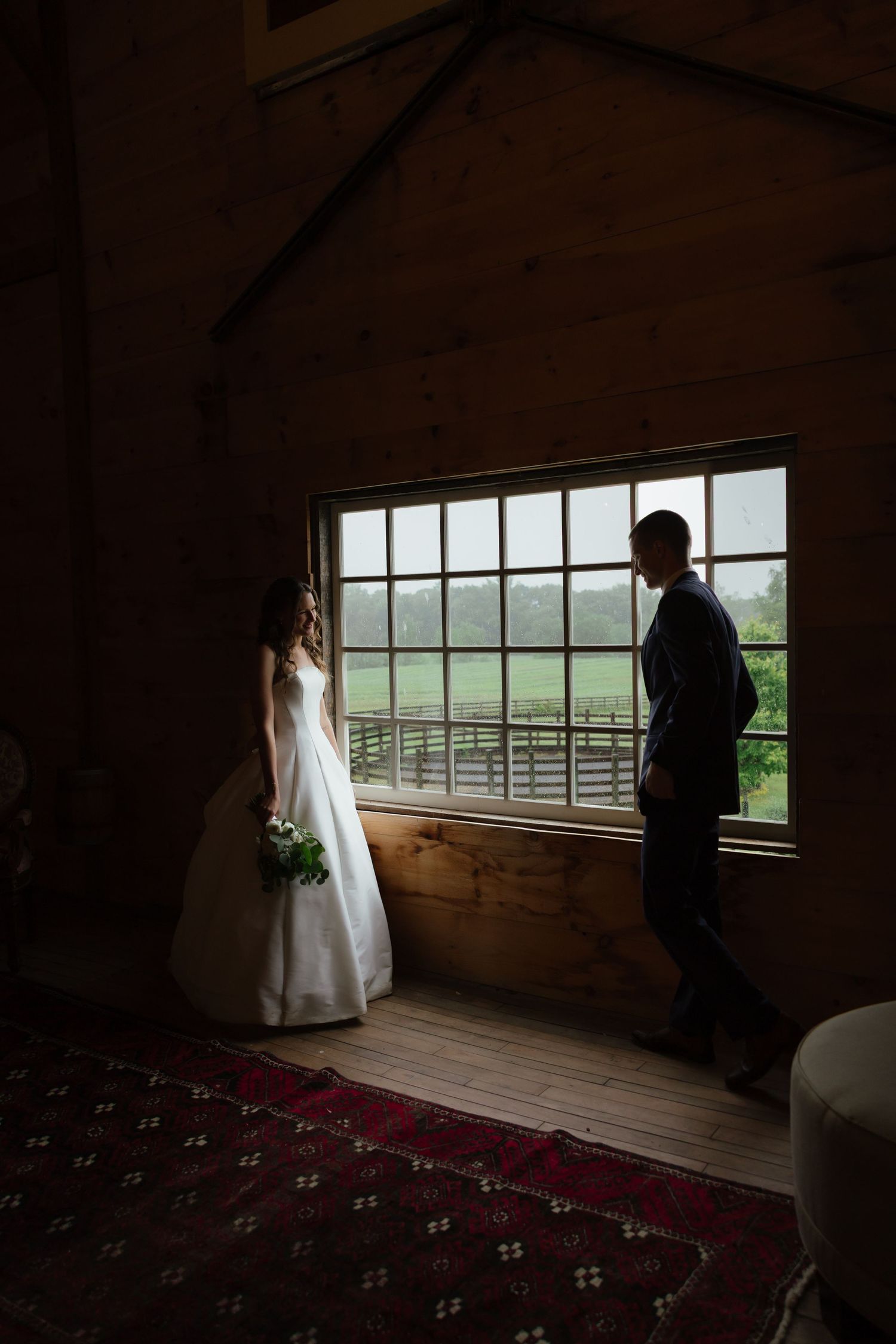 Bride and groom share intimate moment by large barn window with scenic green countryside view.