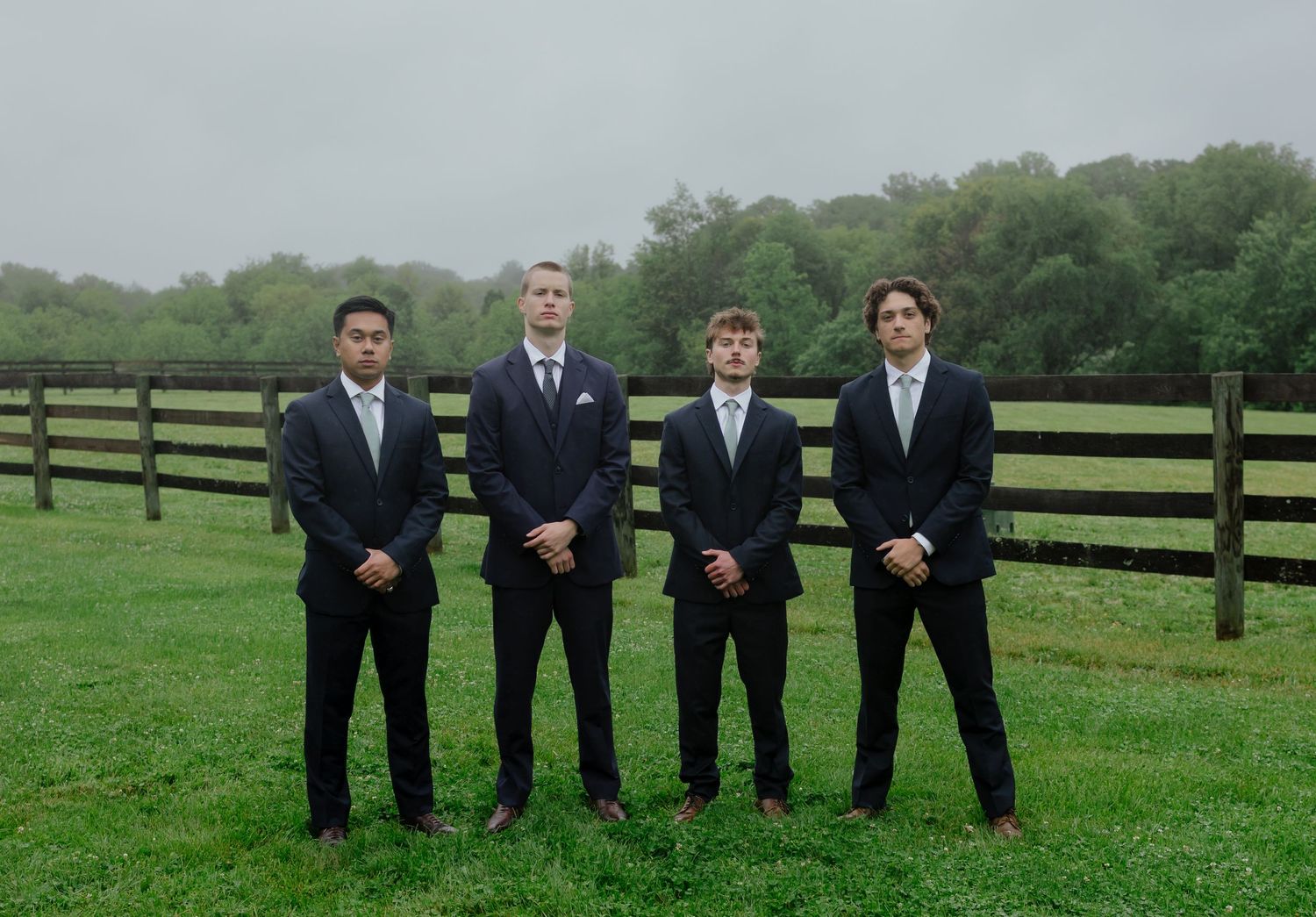 Four groomsmen in dark suits stand in a row on green grass before a wooden fence on overcast day.