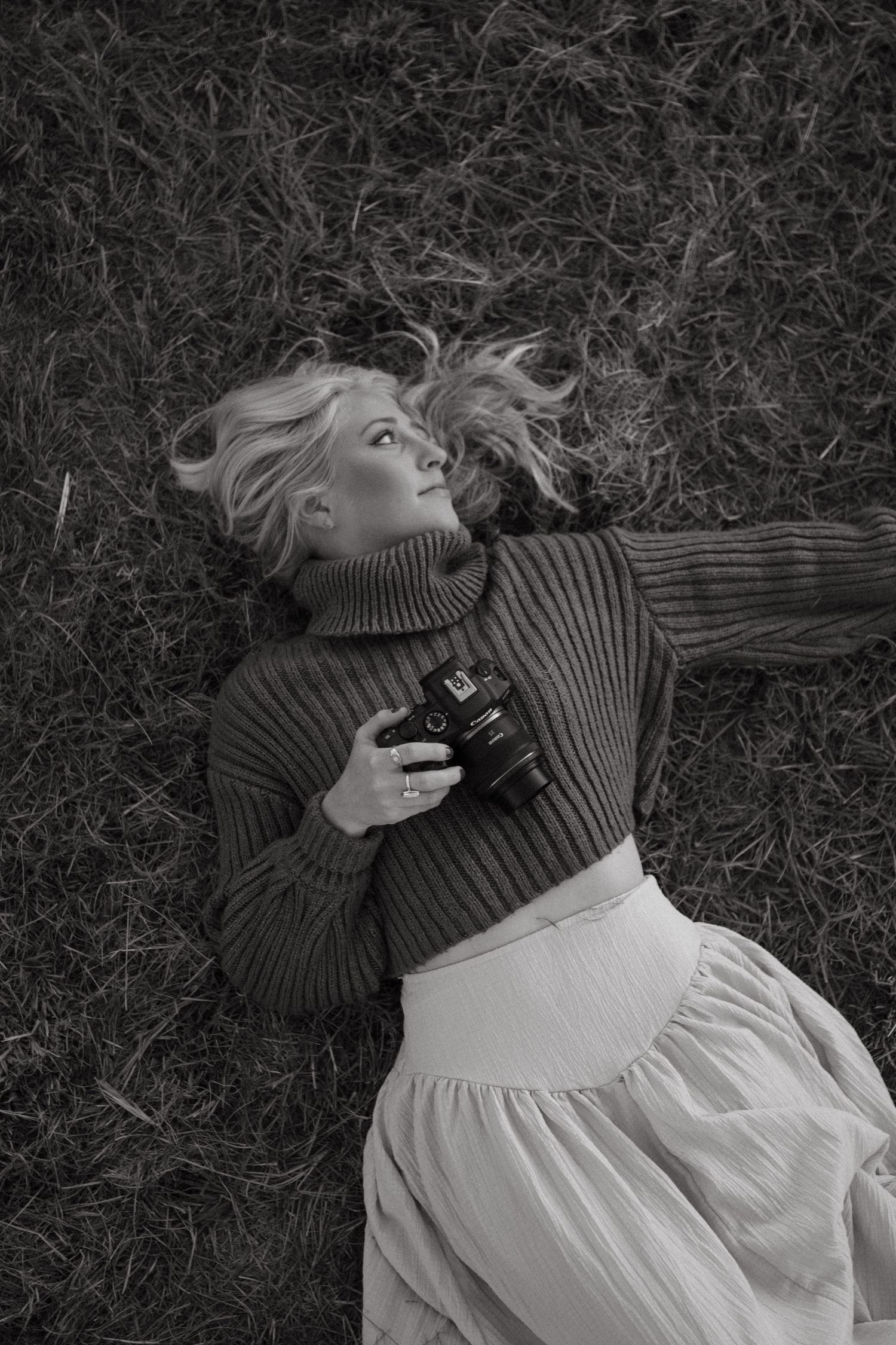 Blonde woman lying on grass holding a camera in a black and white portrait.