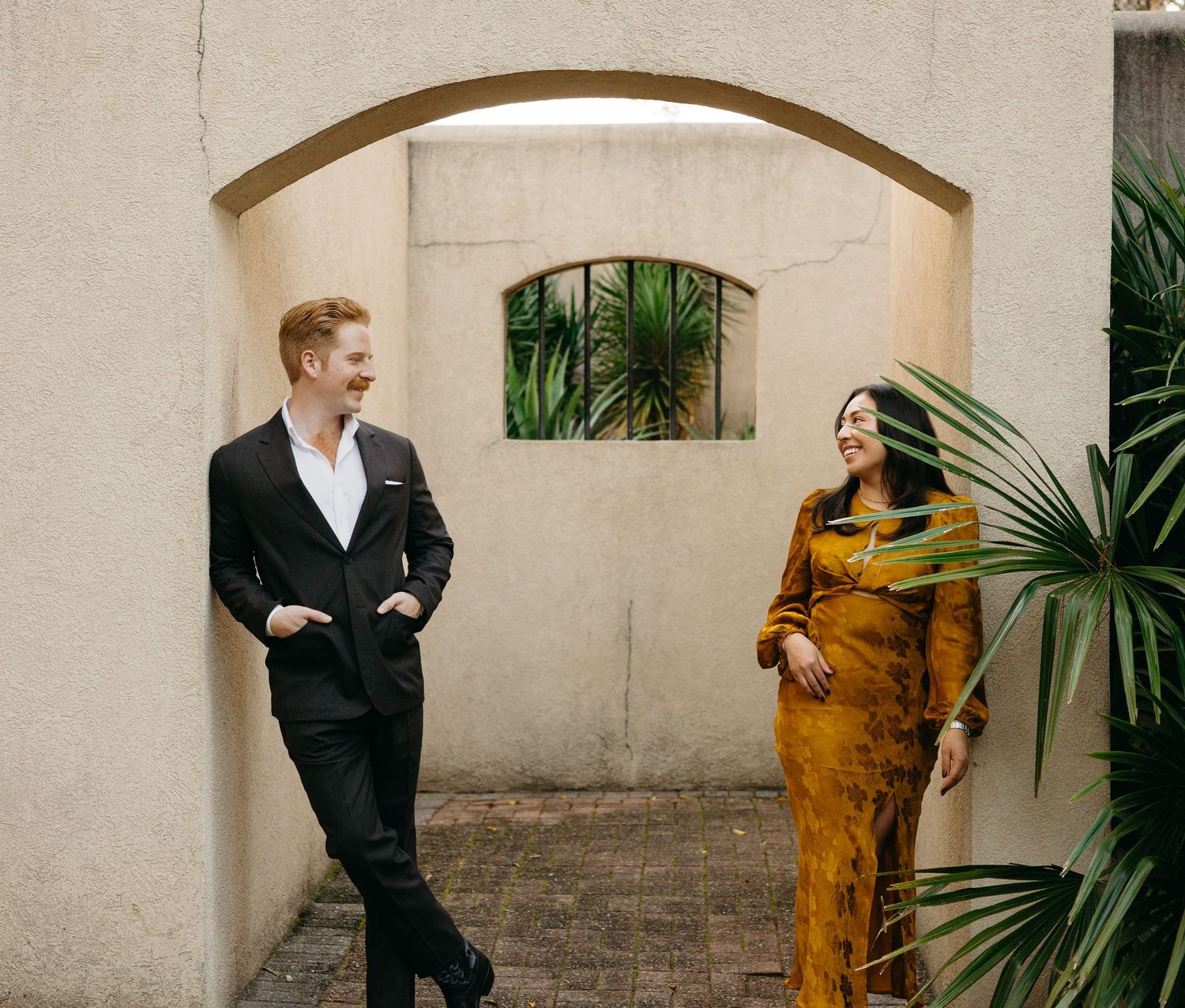 Couple smiling and laughing together near a stucco archway with tropical greenery in warm light.