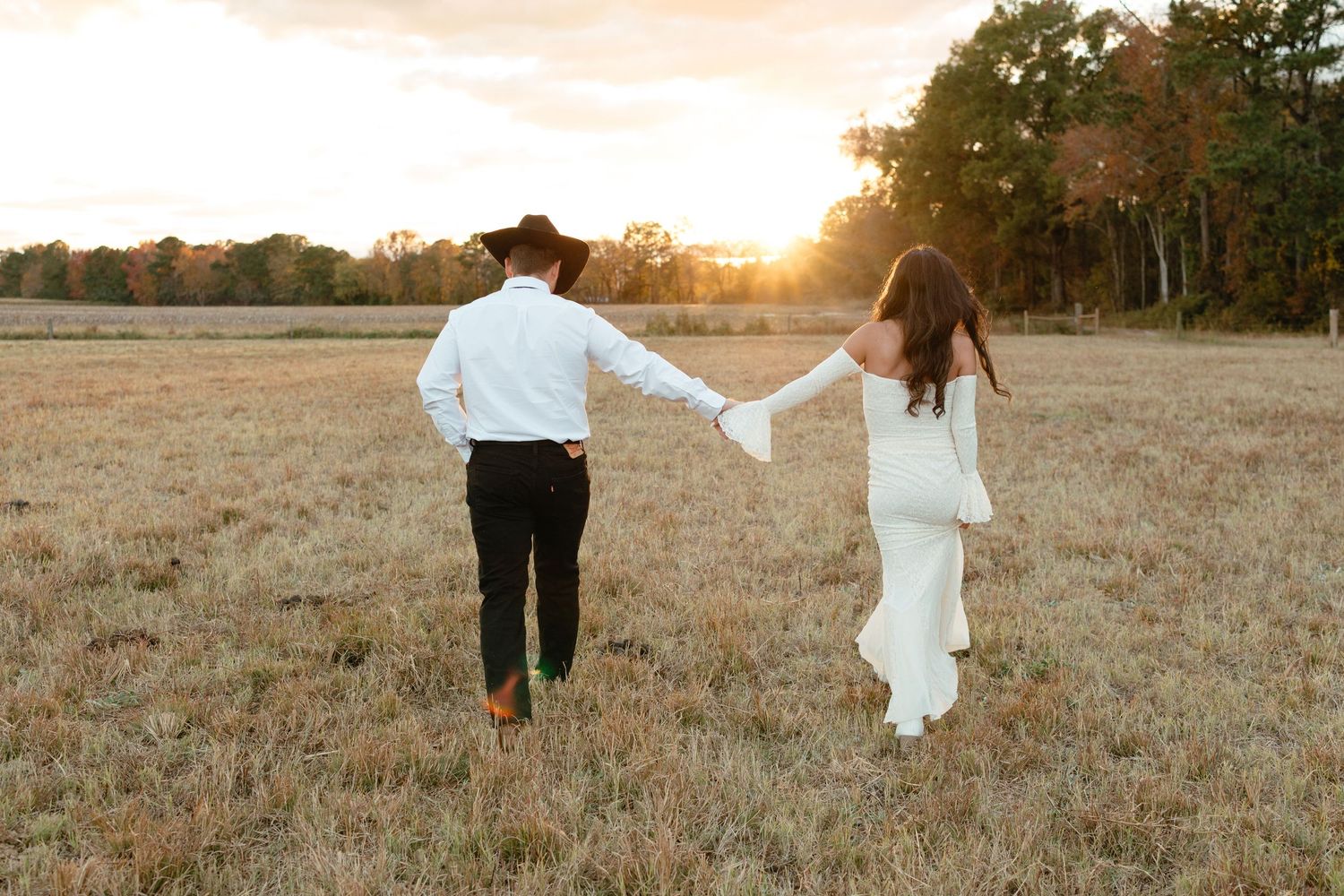 Bride and groom holding hands walking through golden field at sunset in western wedding attire.