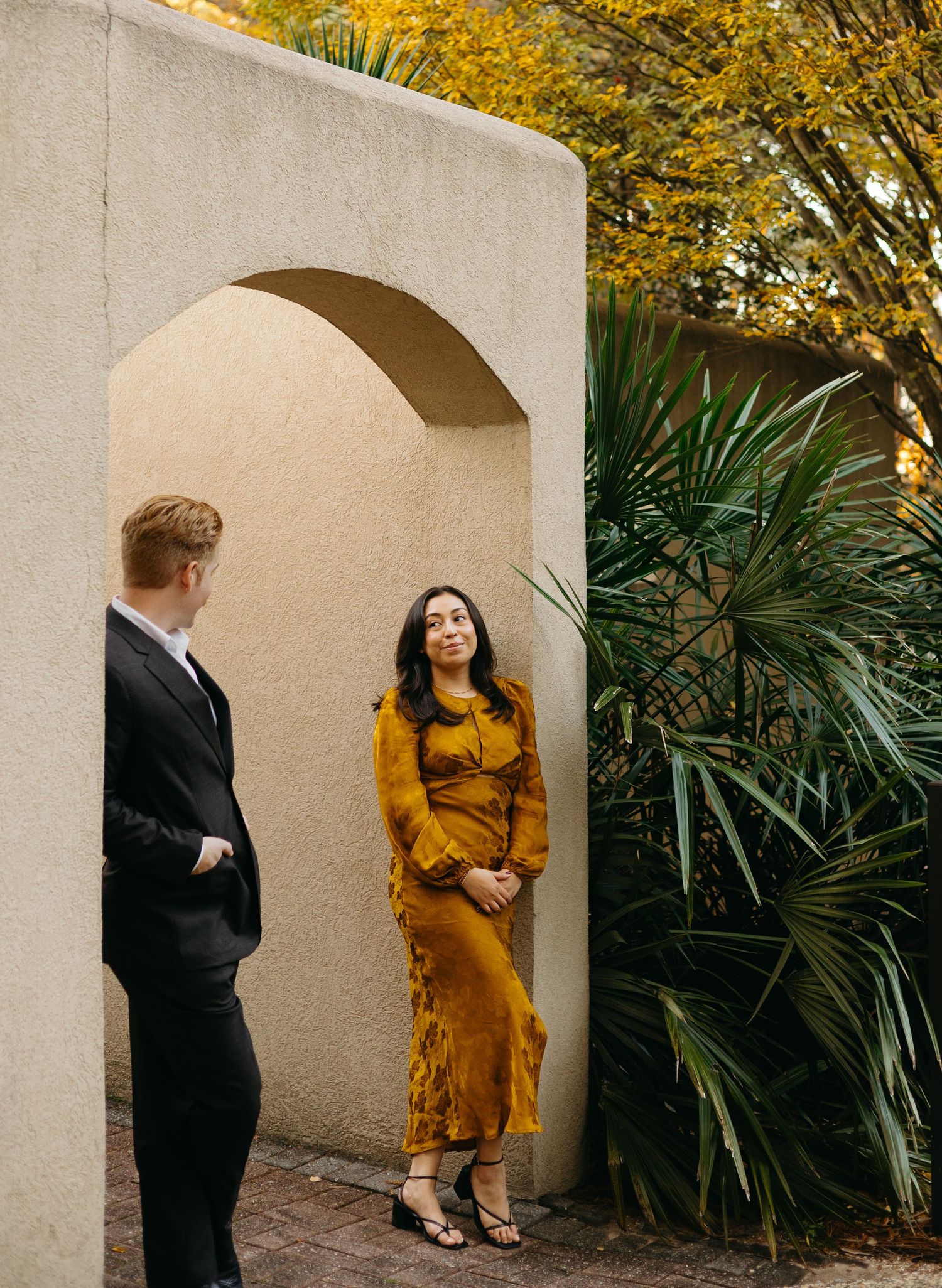 Couple sharing a lighthearted conversation under a stucco archway surrounded by lush tropical plants.