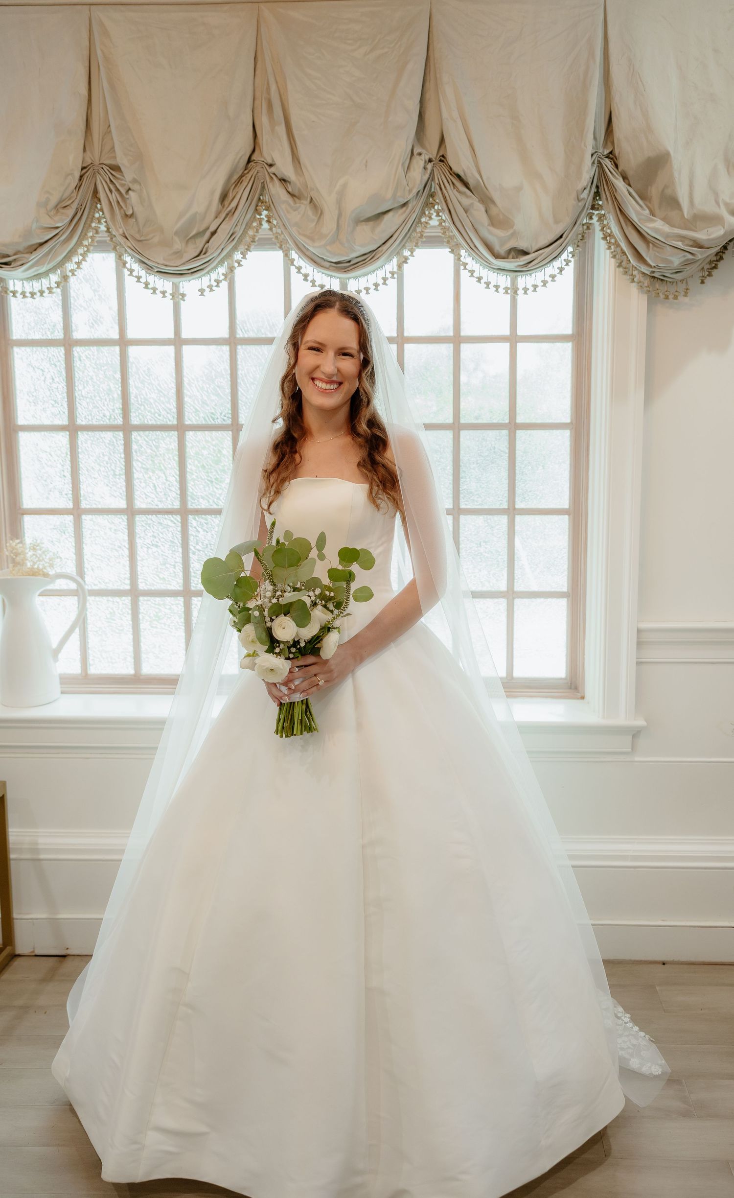 Smiling bride in strapless white ballgown holding floral bouquet standing before elegant draped window.