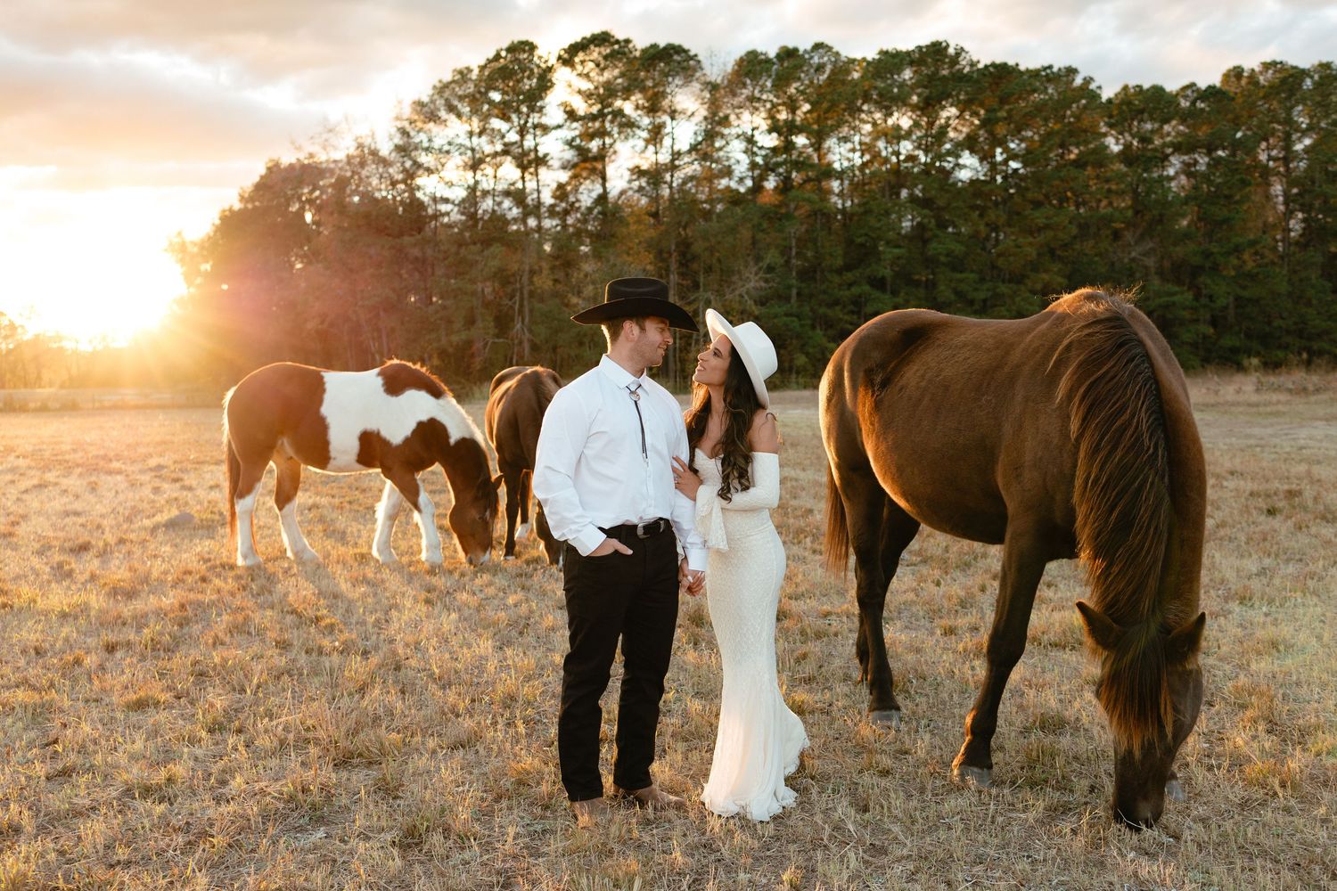 Couple in cowboy hats standing near horses grazing in a sunlit field at golden hour.