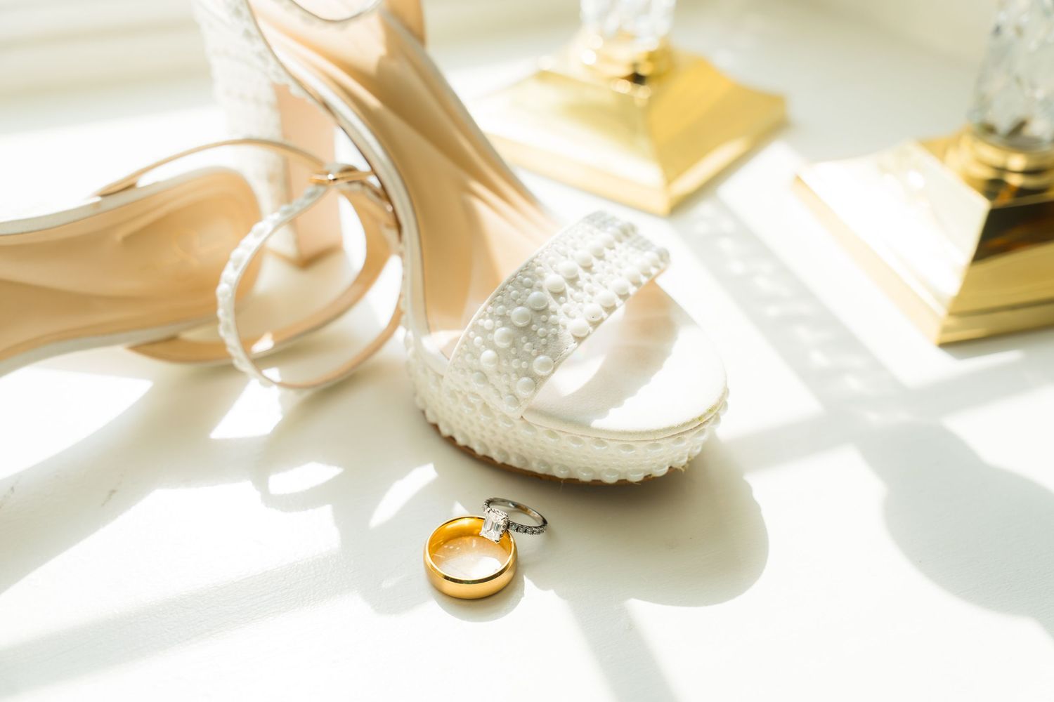 Elegant bridal accessories including crystal heels, gold rings, and a golden clutch on a white surface.