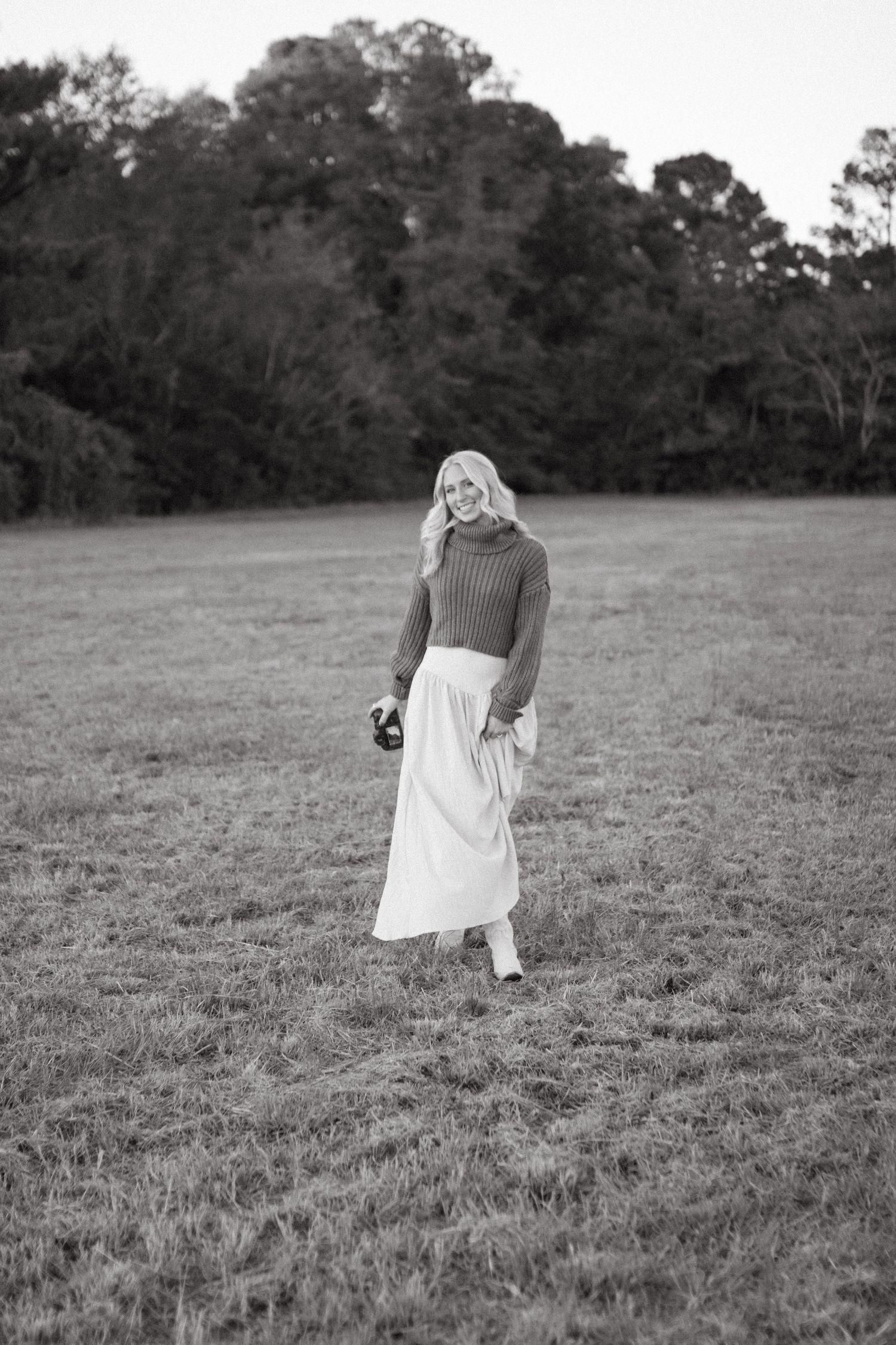 A blonde woman in a knit sweater and white maxi skirt stands in a grassy field holding a camera.