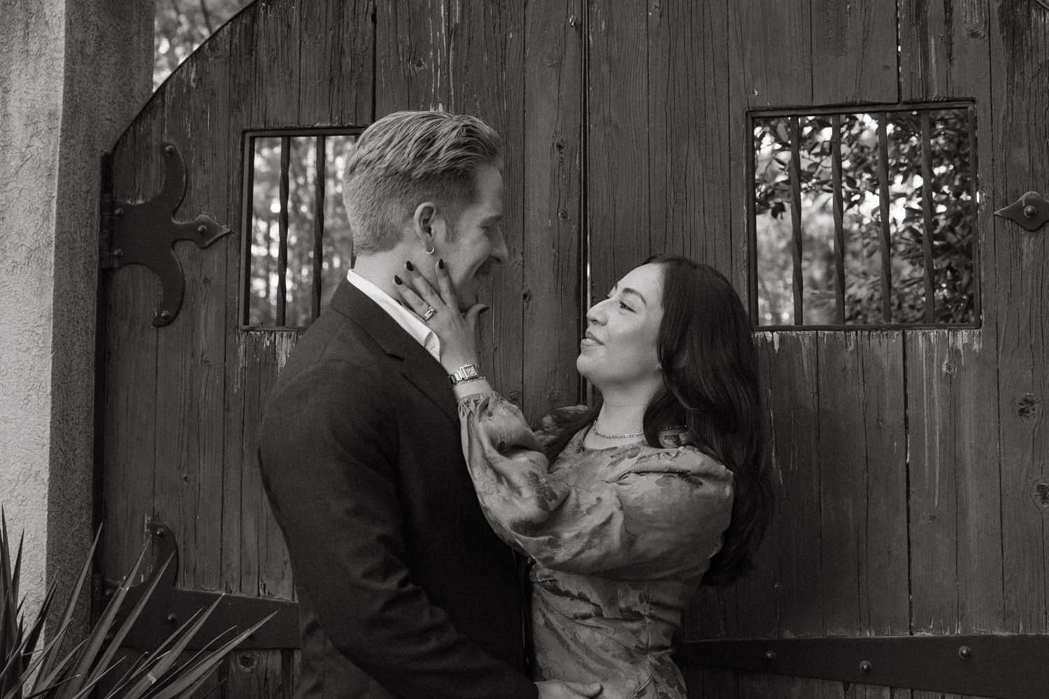 Black and white photo of couple nearly kissing before rustic wooden gate, woman's hand tenderly on man's face.