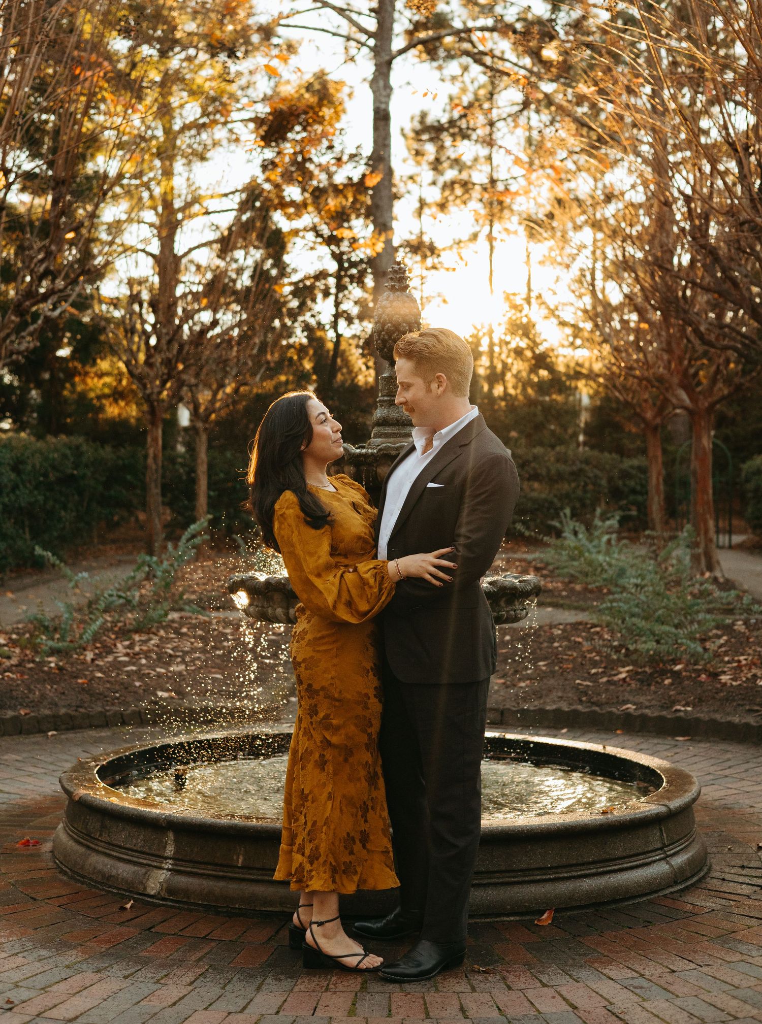 Couple embracing near a circular fountain at sunset in an autumn garden.