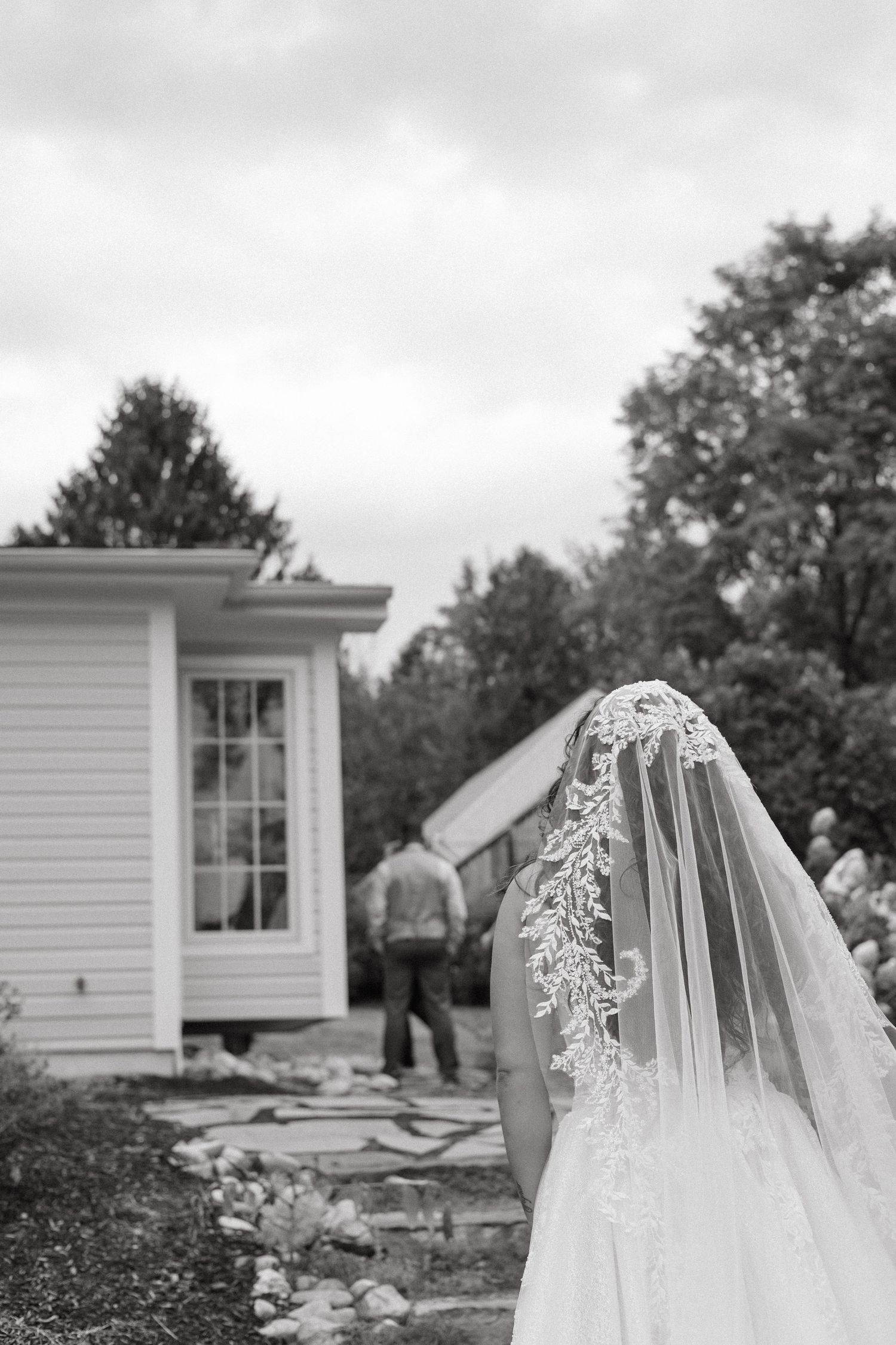 Black and white photo of a bride in lace veil standing outdoors near a white building with a man walking in background.