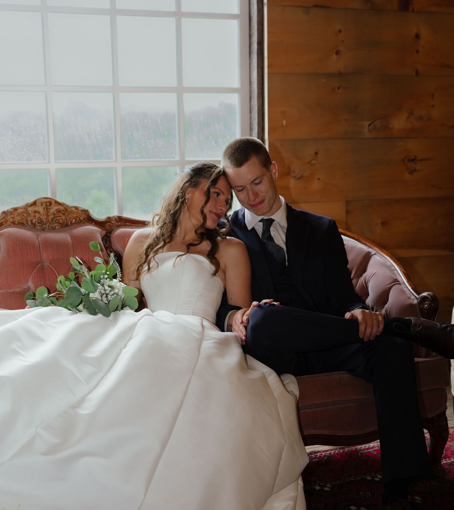 Bride and groom sit together on a vintage sofa in a cozy rustic barn wedding venue.
