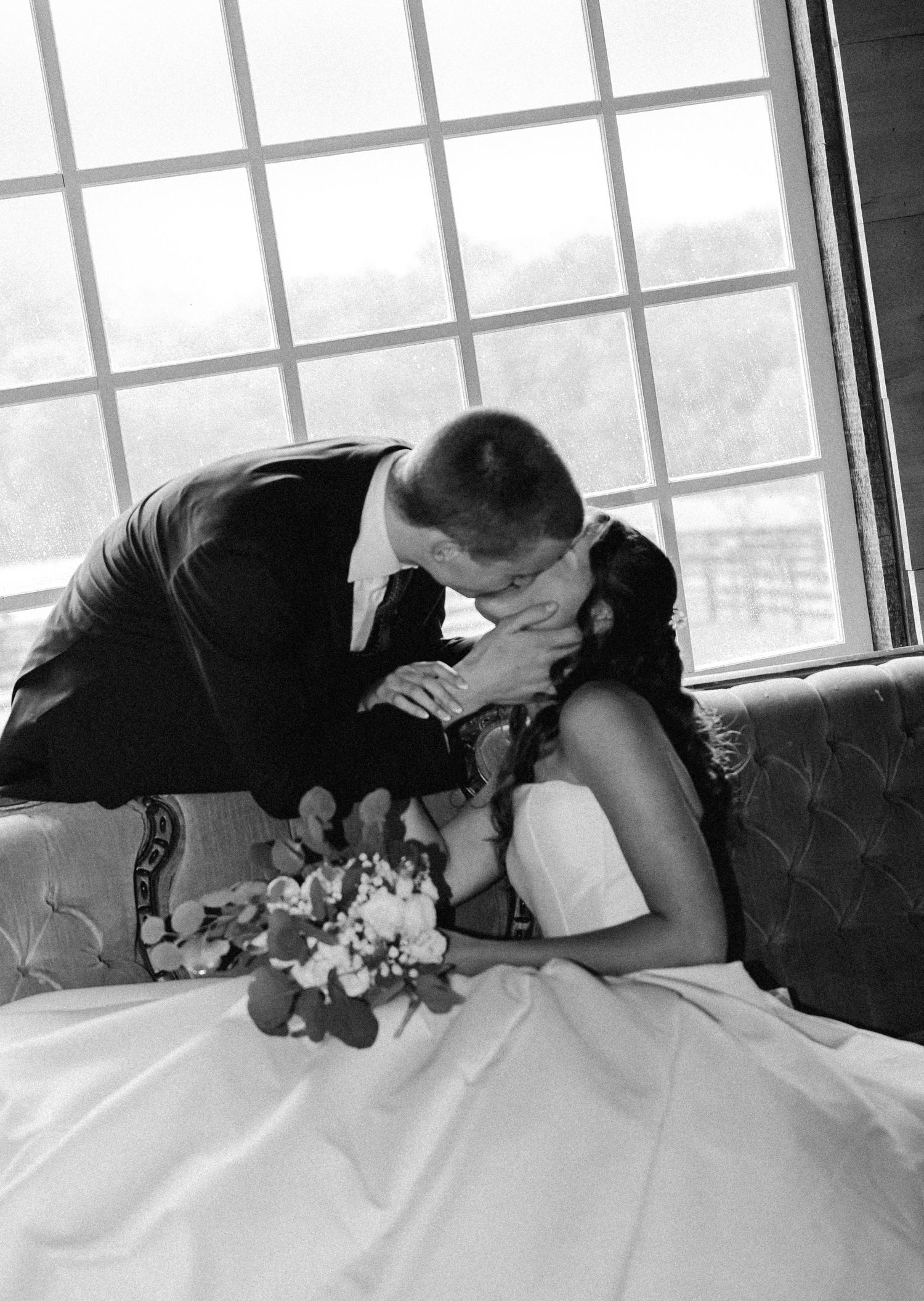 Black and white photo of groom tenderly kissing bride holding bouquet on vintage sofa by window.