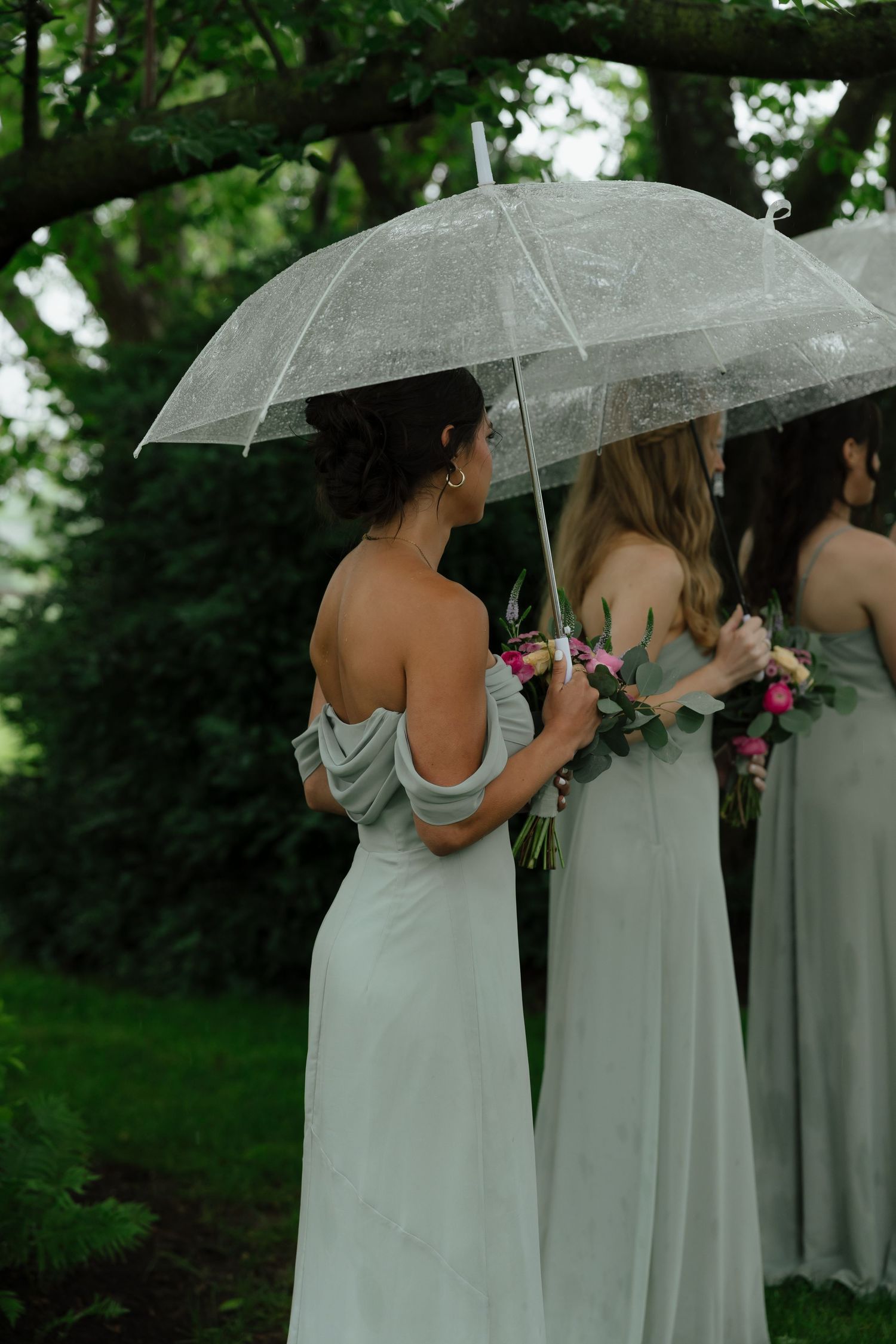 Bridesmaids in sage green off-shoulder gowns holding bouquets under clear umbrellas in garden.
