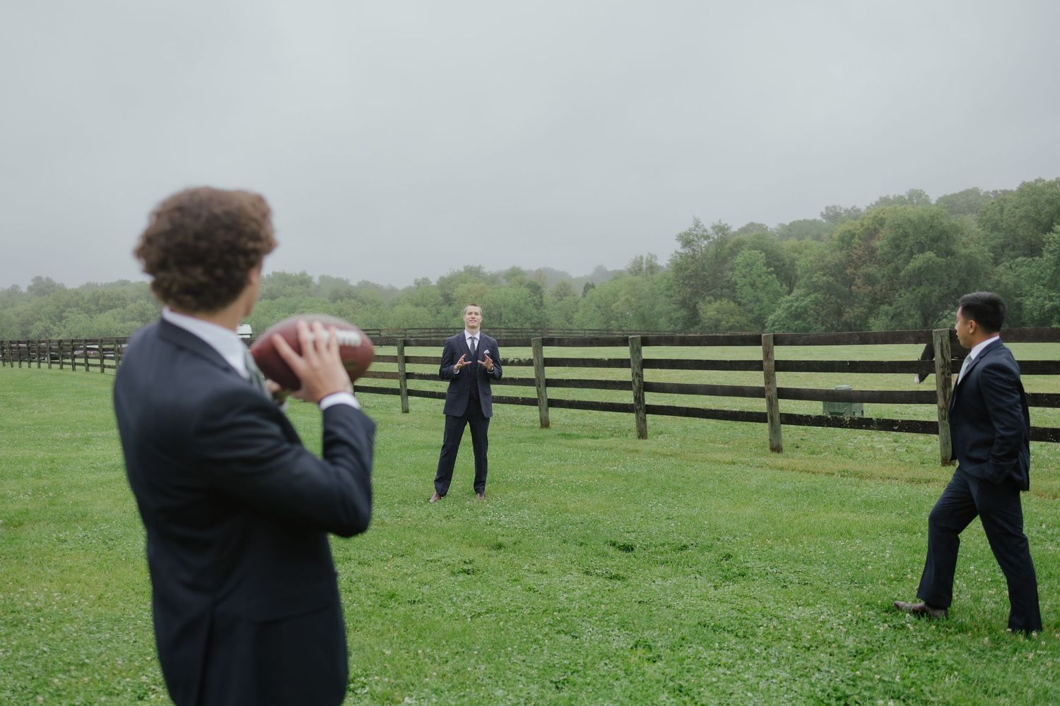 Groomsmen in dark suits toss a football on green farm field with wooden fence in background.