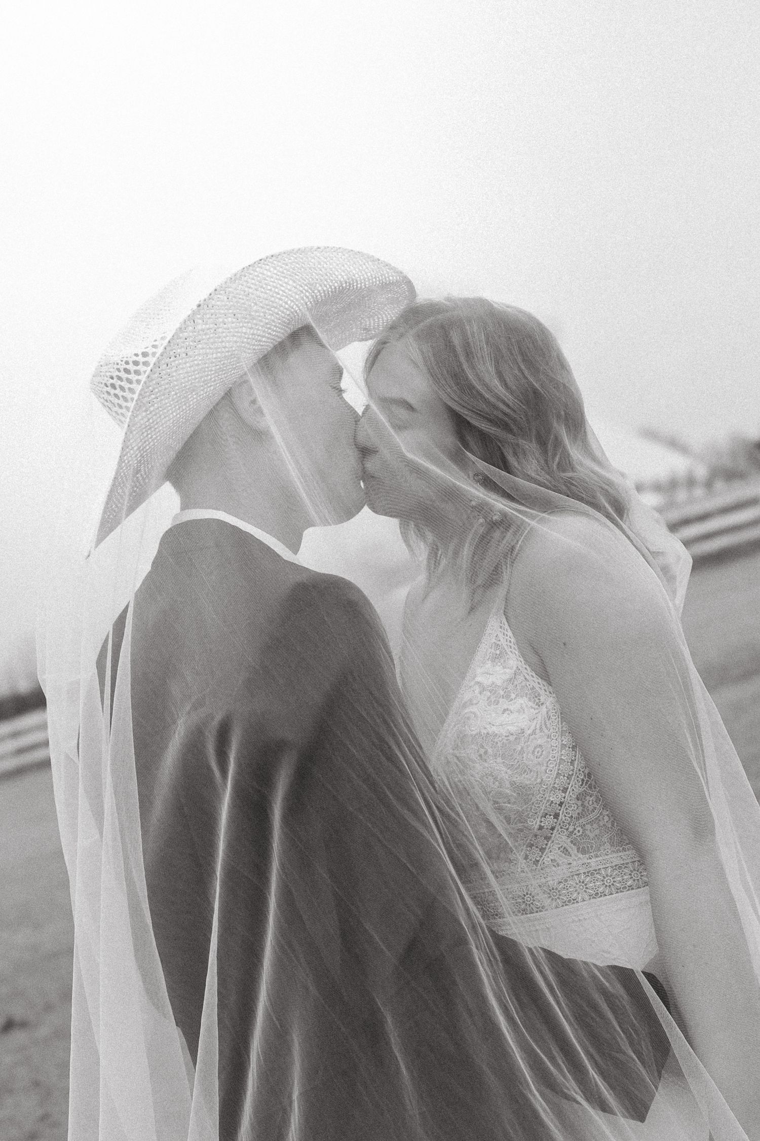 Black and white romantic photo of bride and groom kissing under a flowing veil outdoors.