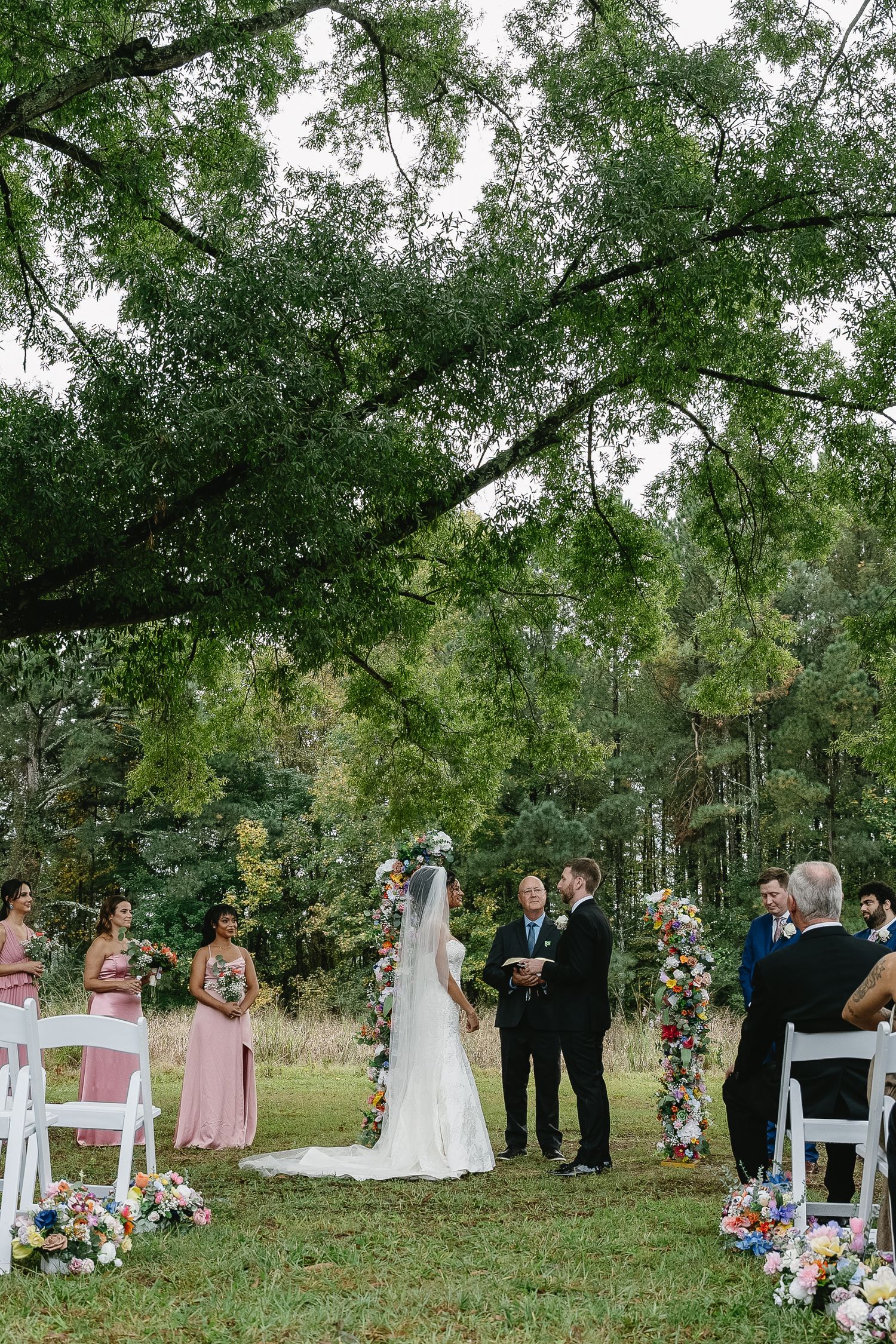 Outdoor wedding ceremony under a large oak tree with colorful floral arch and white chairs.