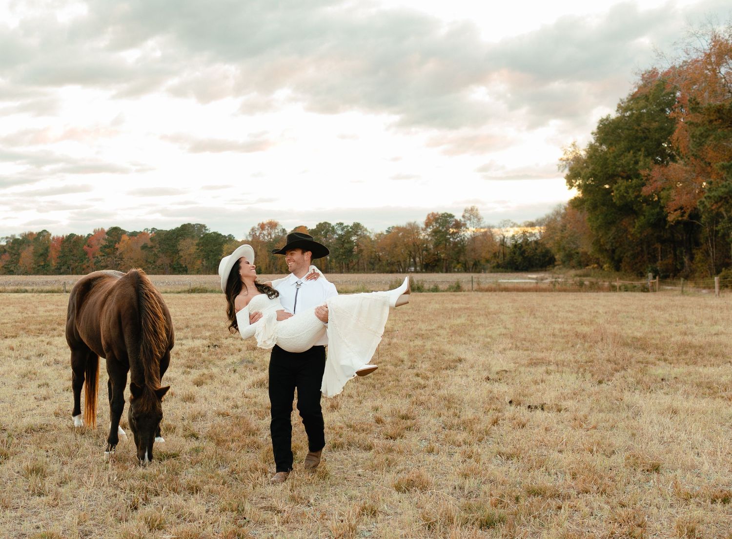 Groom carrying bride in white dress beside grazing horse in open autumn field.
