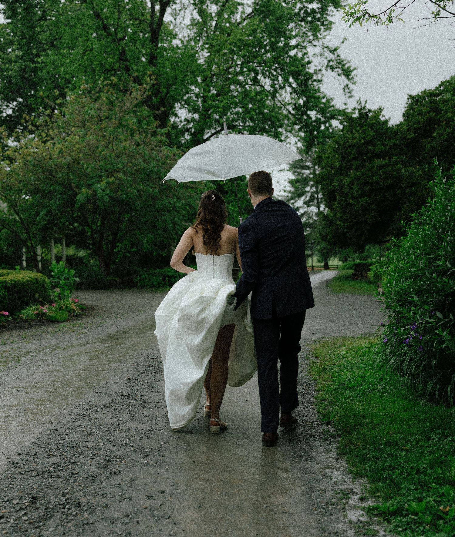 Bride and groom walking hand in hand down a rainy garden path sharing a white umbrella on their wedding day.
