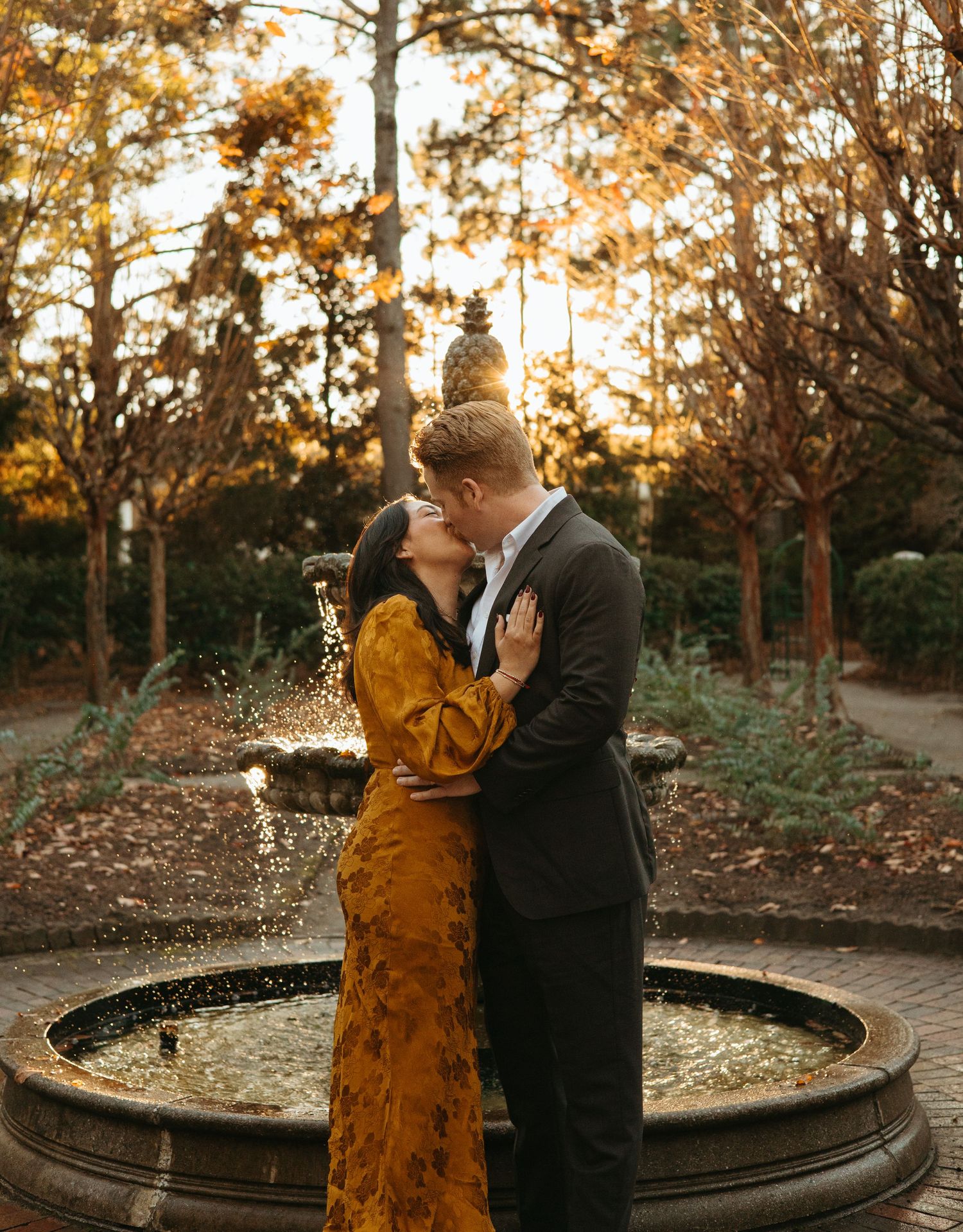 Romantic couple sharing a kiss by a garden fountain at golden hour, woman in mustard yellow dress.
