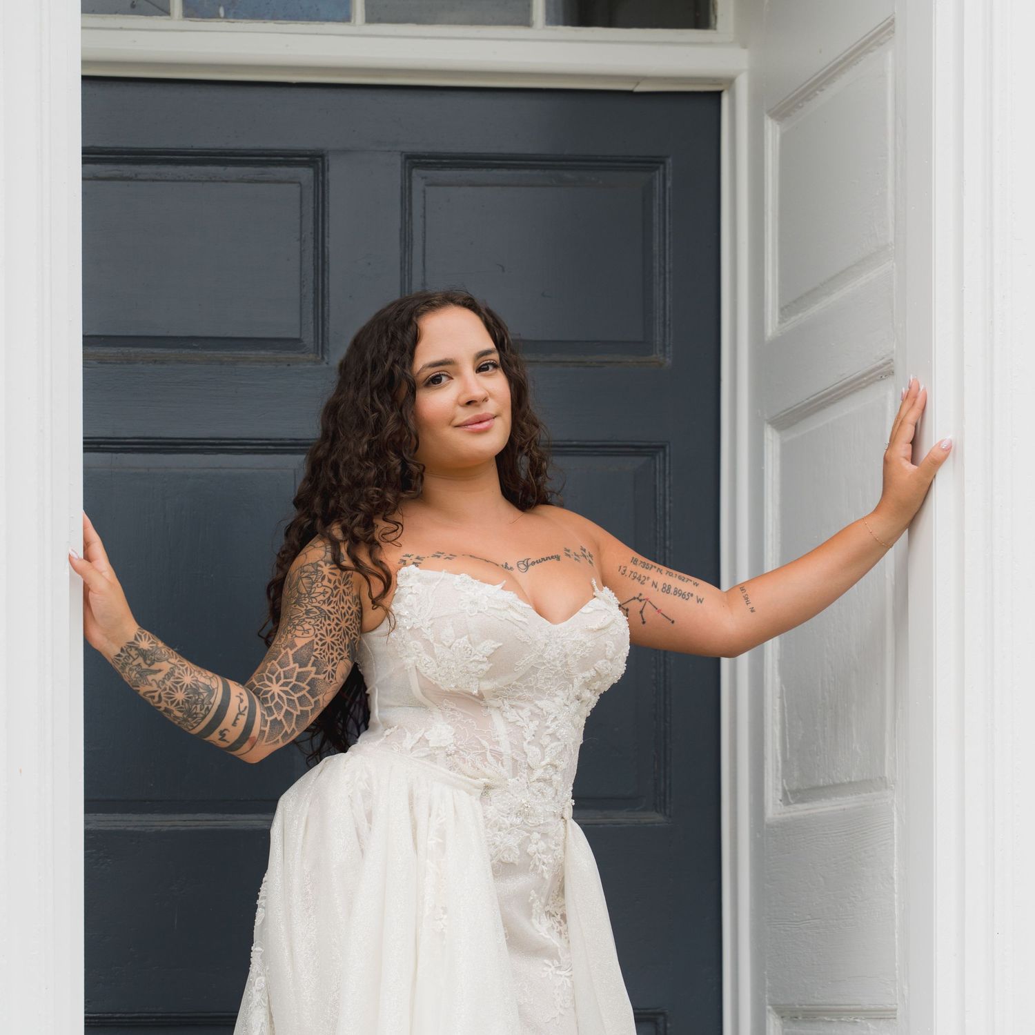 Tattooed bride in strapless lace wedding dress poses in doorway with arms outstretched smiling.