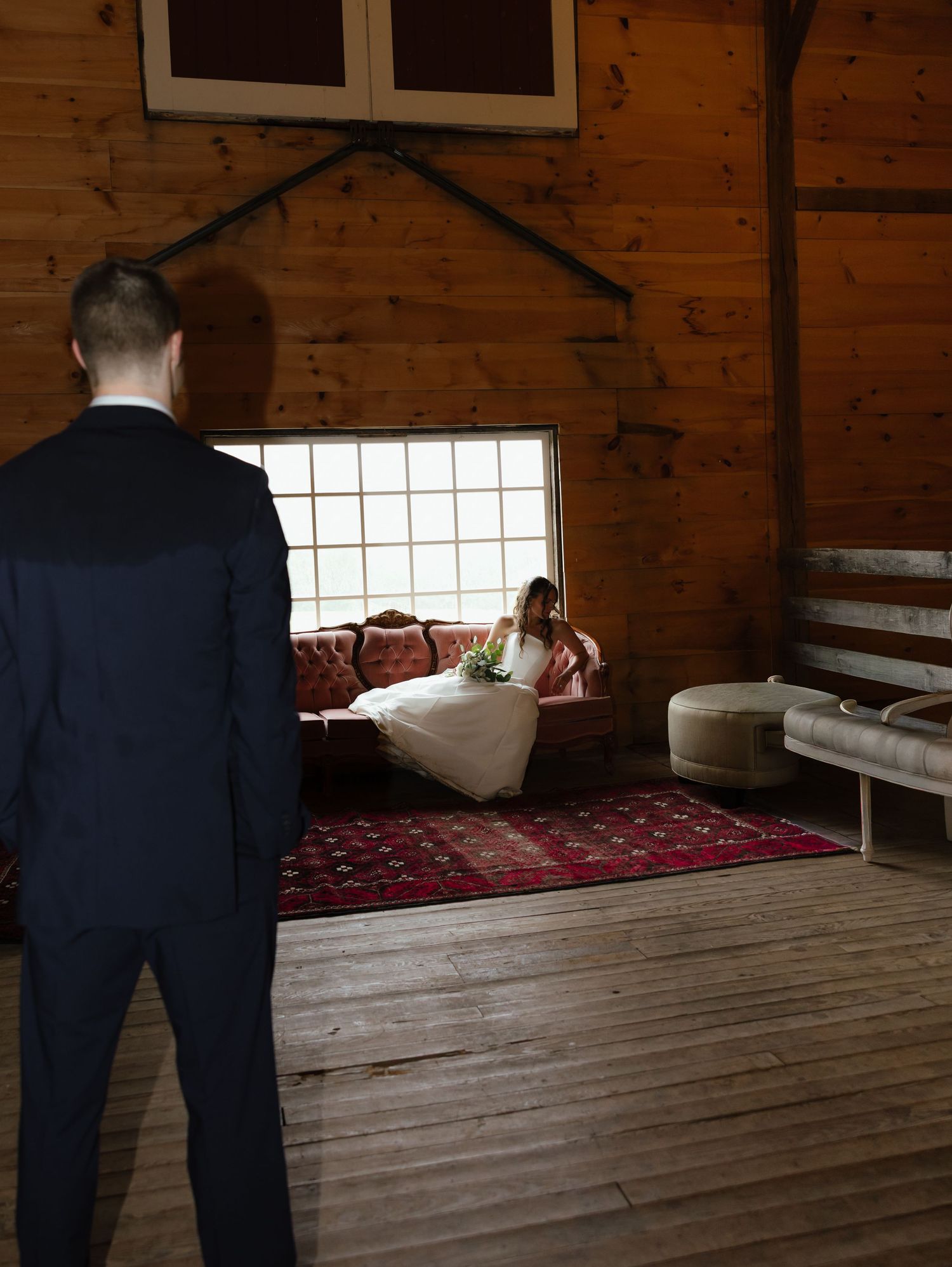 Groom in navy suit watches bride in white dress seated by window inside rustic wooden barn.