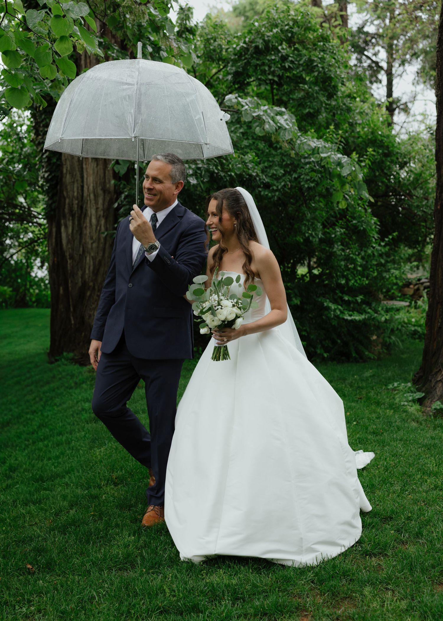 Father walks bride down the aisle under a clear umbrella in a lush green garden.