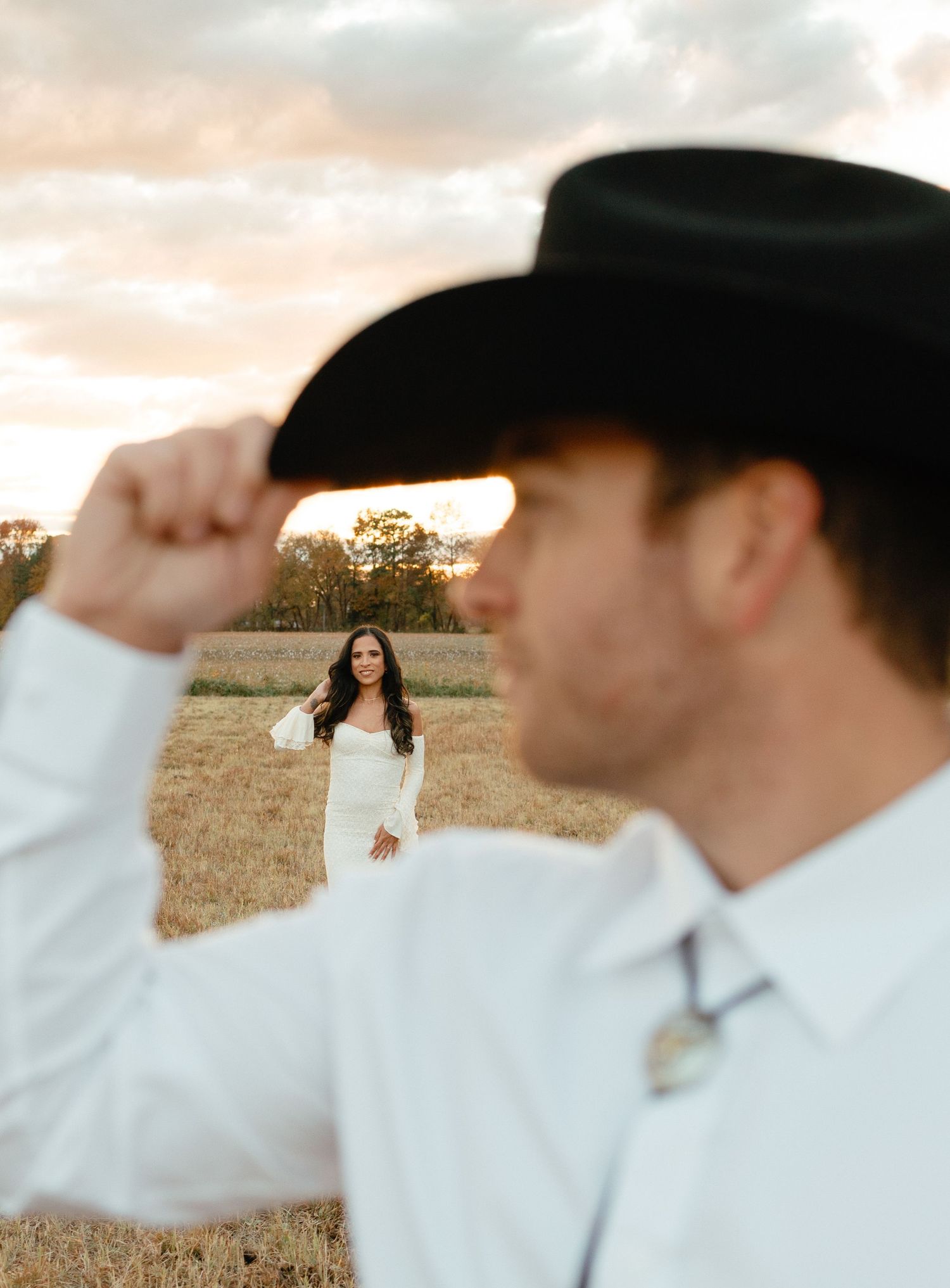 Groom in black cowboy hat tips his brim while bride in white gown stands in a sun-drenched field behind him.