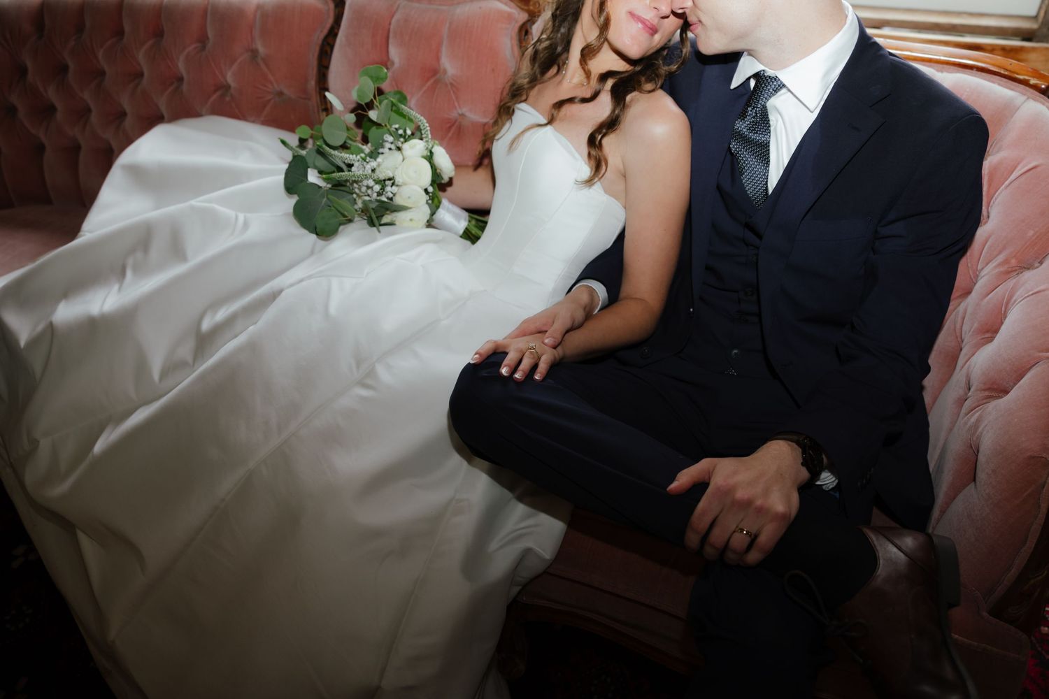Close-up of bride and groom holding hands while seated together on a vintage pink velvet sofa.