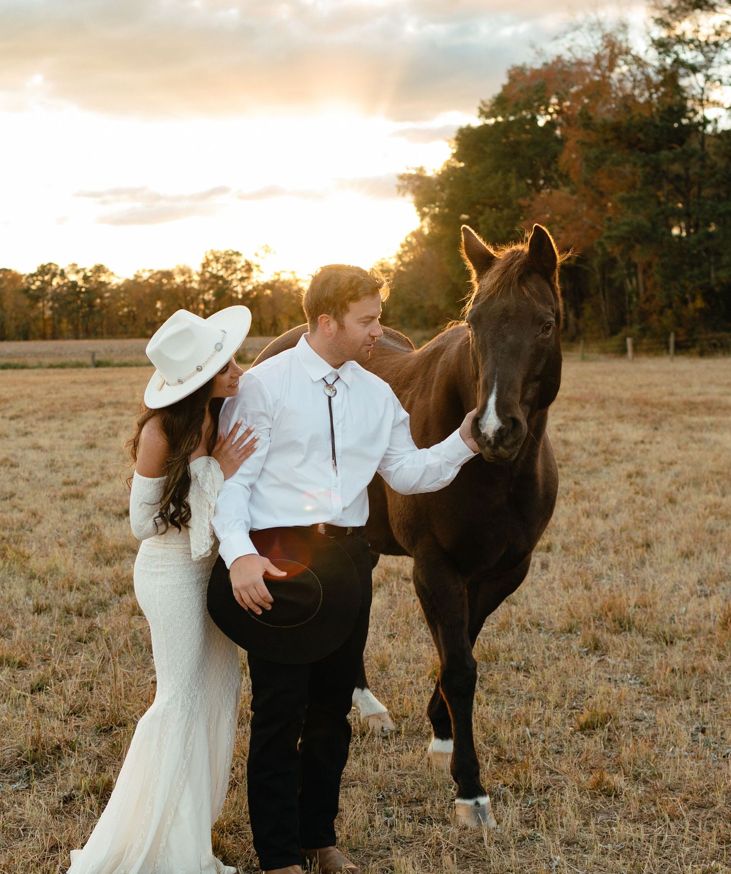 Couple in western attire standing with a brown horse in a golden field at sunset.