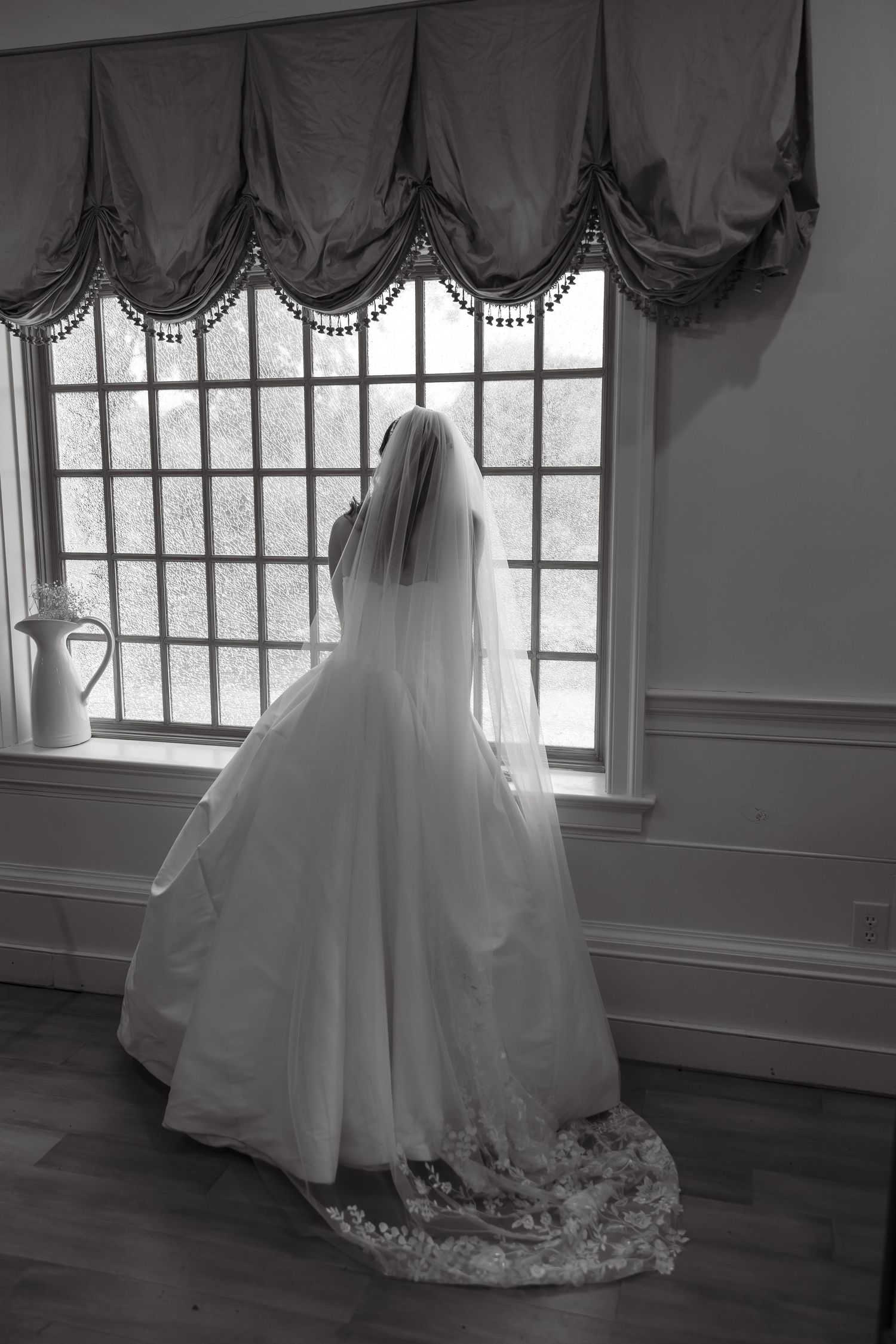 Bride in full ball gown with lace-trimmed veil gazes out large window in black and white portrait.