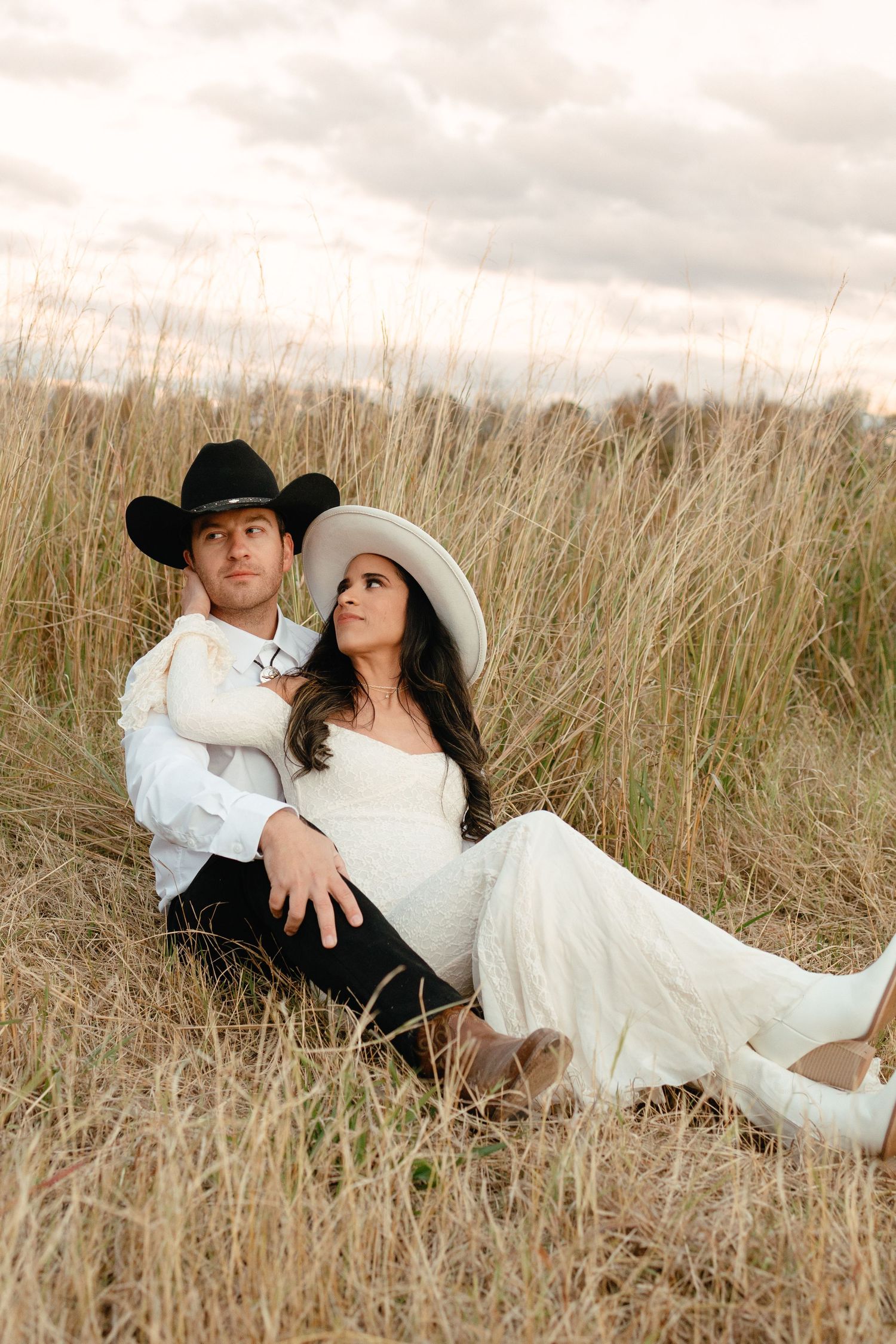 Bride and groom in cowboy hats sitting in tall golden grass field during romantic outdoor photoshoot.
