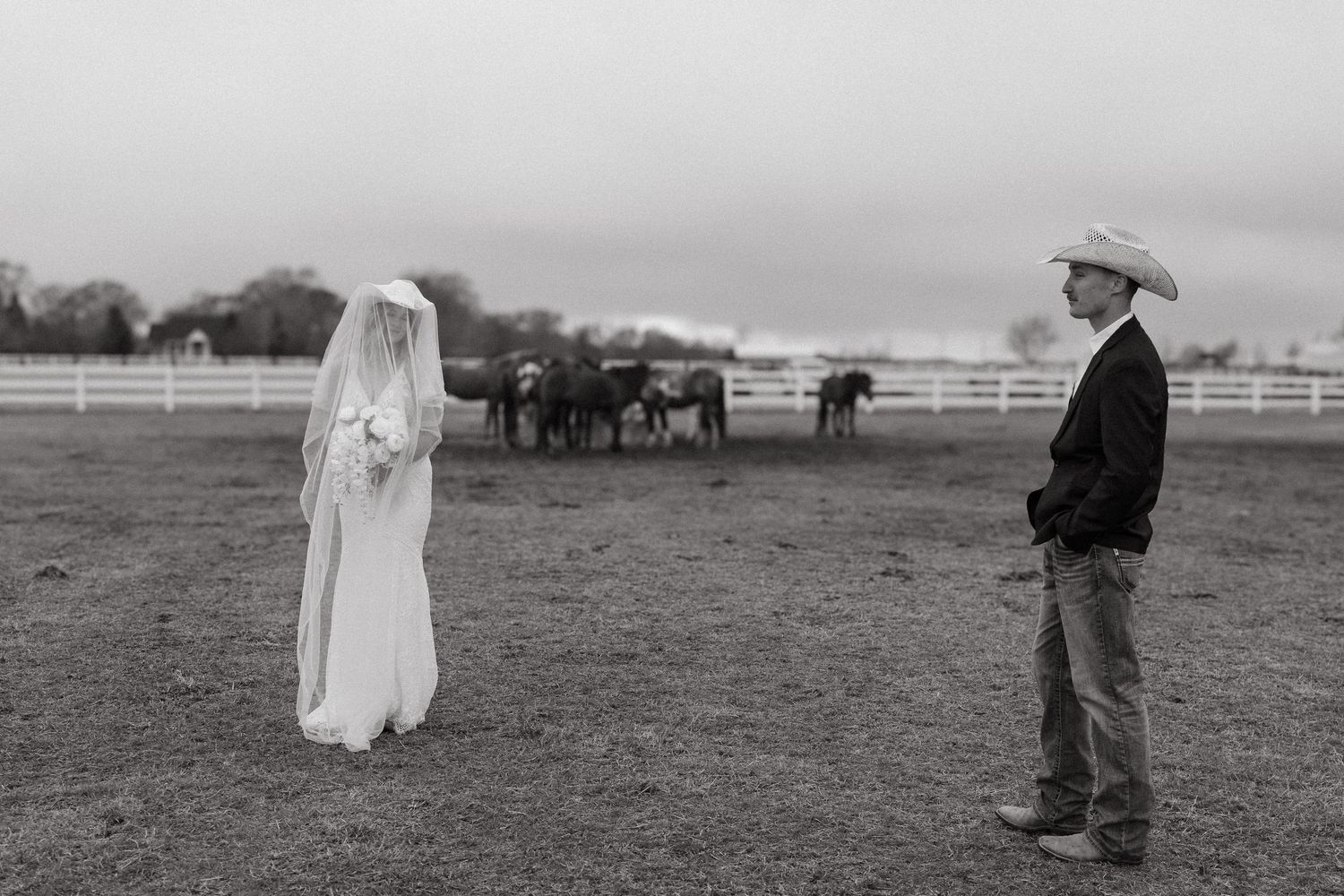 Black and white photo of bride in white gown and veiled cowboy groom in hat standing in ranch field with horses.