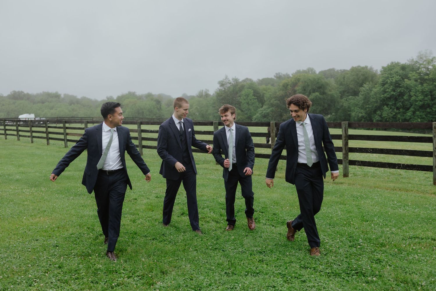 Four groomsmen in dark suits walking and laughing together on a misty farm field.