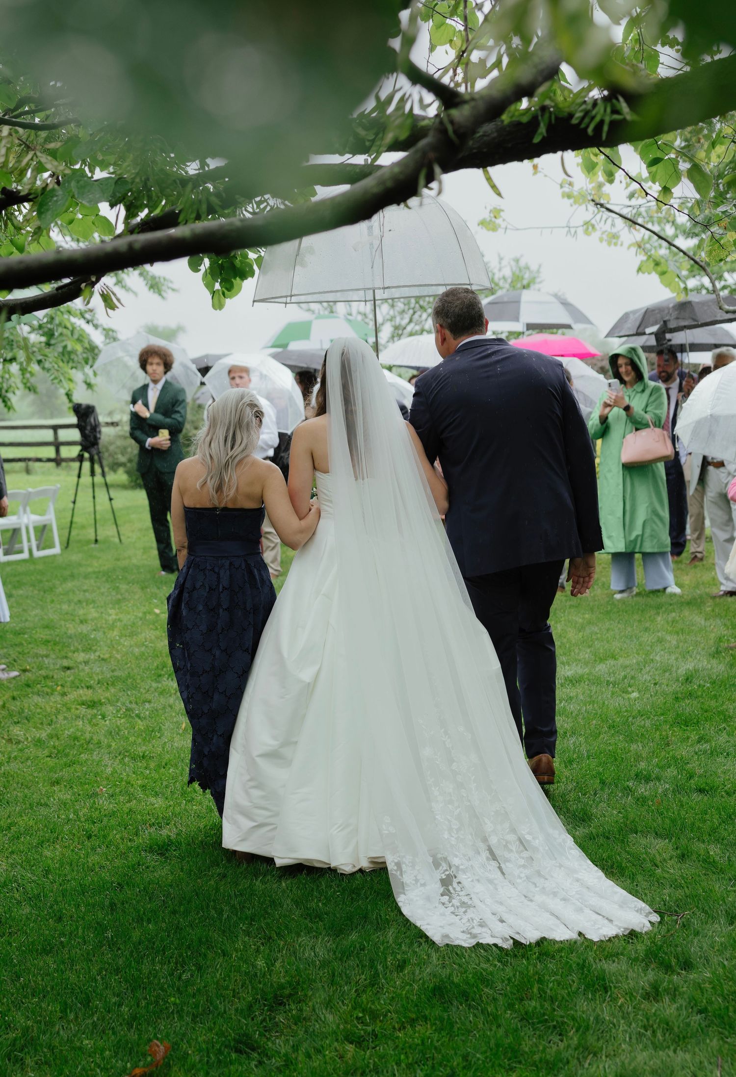 Bride escorted down aisle by parents during outdoor rainy wedding ceremony with umbrella-holding guests.