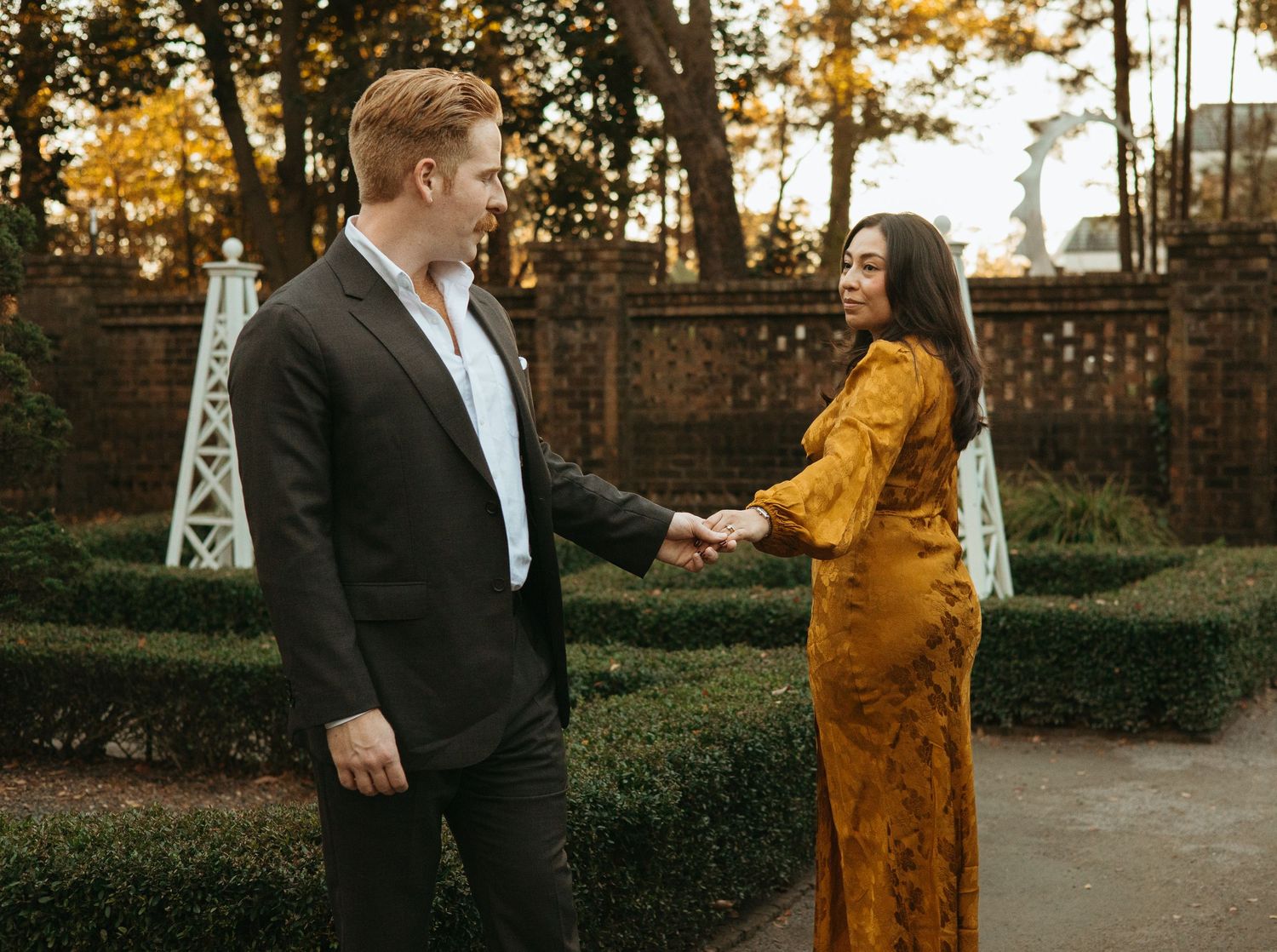 Couple holding hands smiling at each other in garden with trimmed boxwood hedges backdrop.