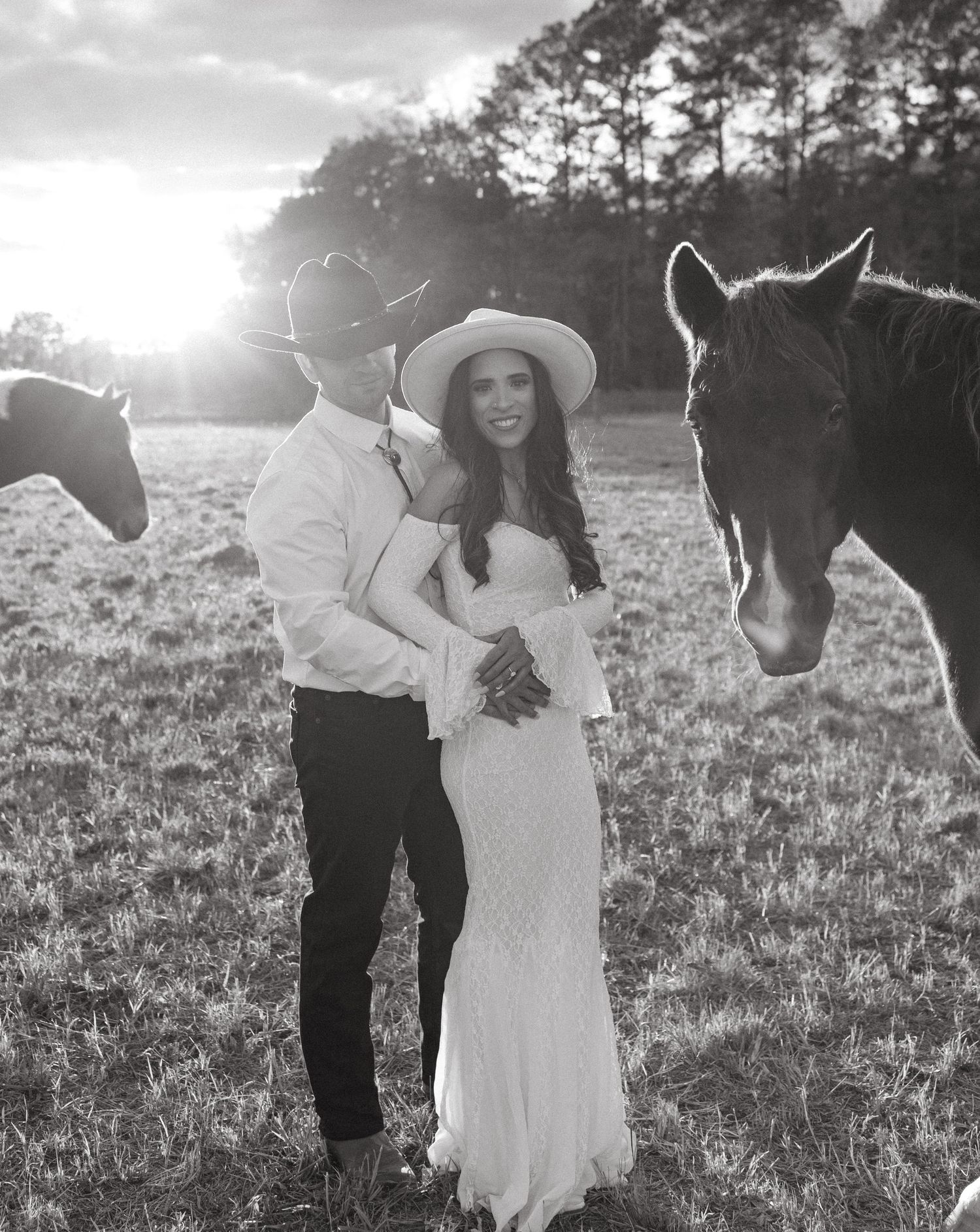 Black and white photo of a couple in cowboy hats embracing in a field with horses at sunset.