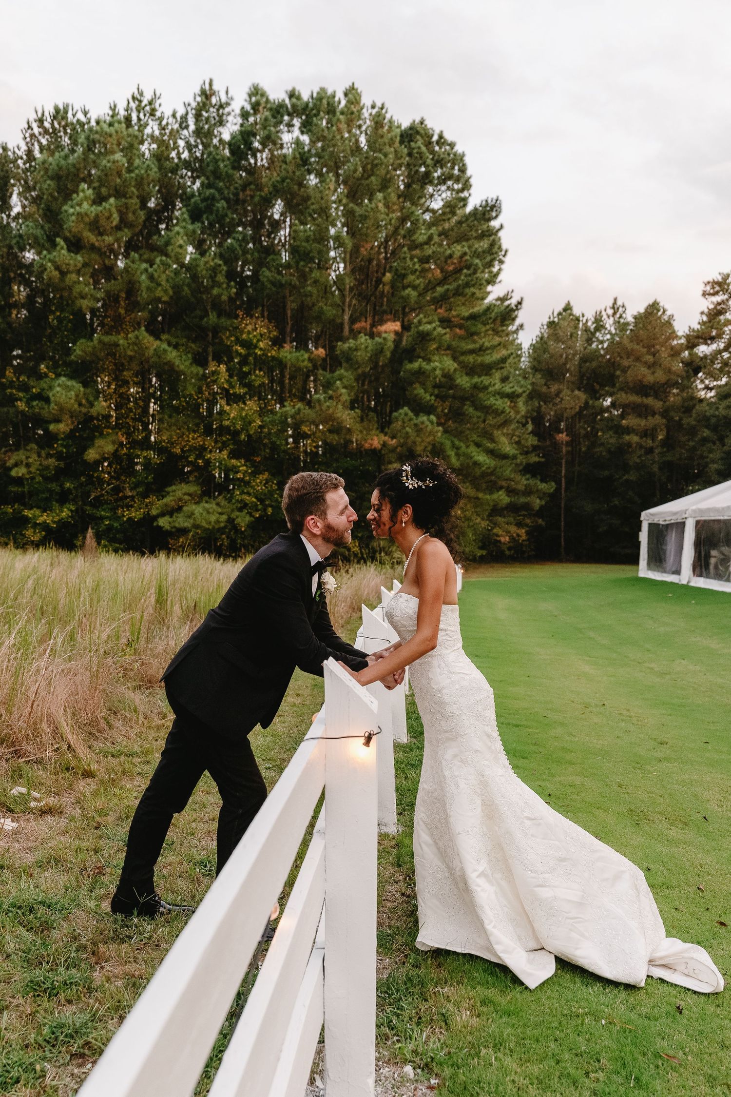 Bride and groom share a romantic moment leaning against a white fence with lush green trees in the background.