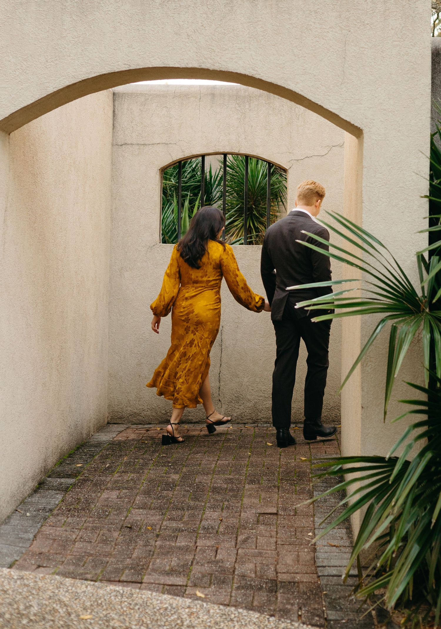 Couple walking hand in hand through a stucco archway surrounded by tropical palm plants.