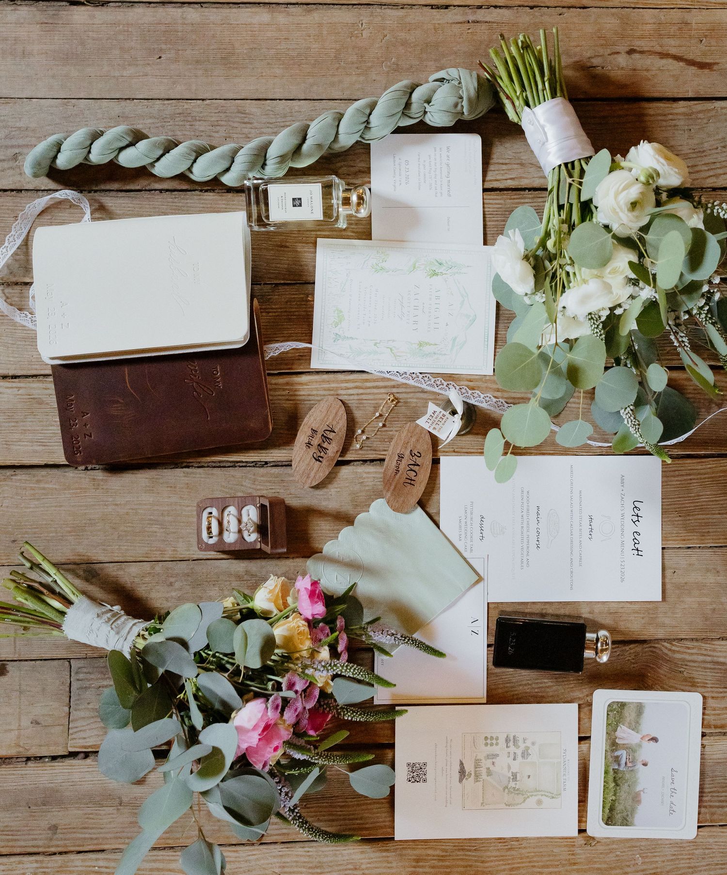 Flat lay of wedding stationery, eucalyptus bouquet, rings, perfume, and accessories on rustic wood.