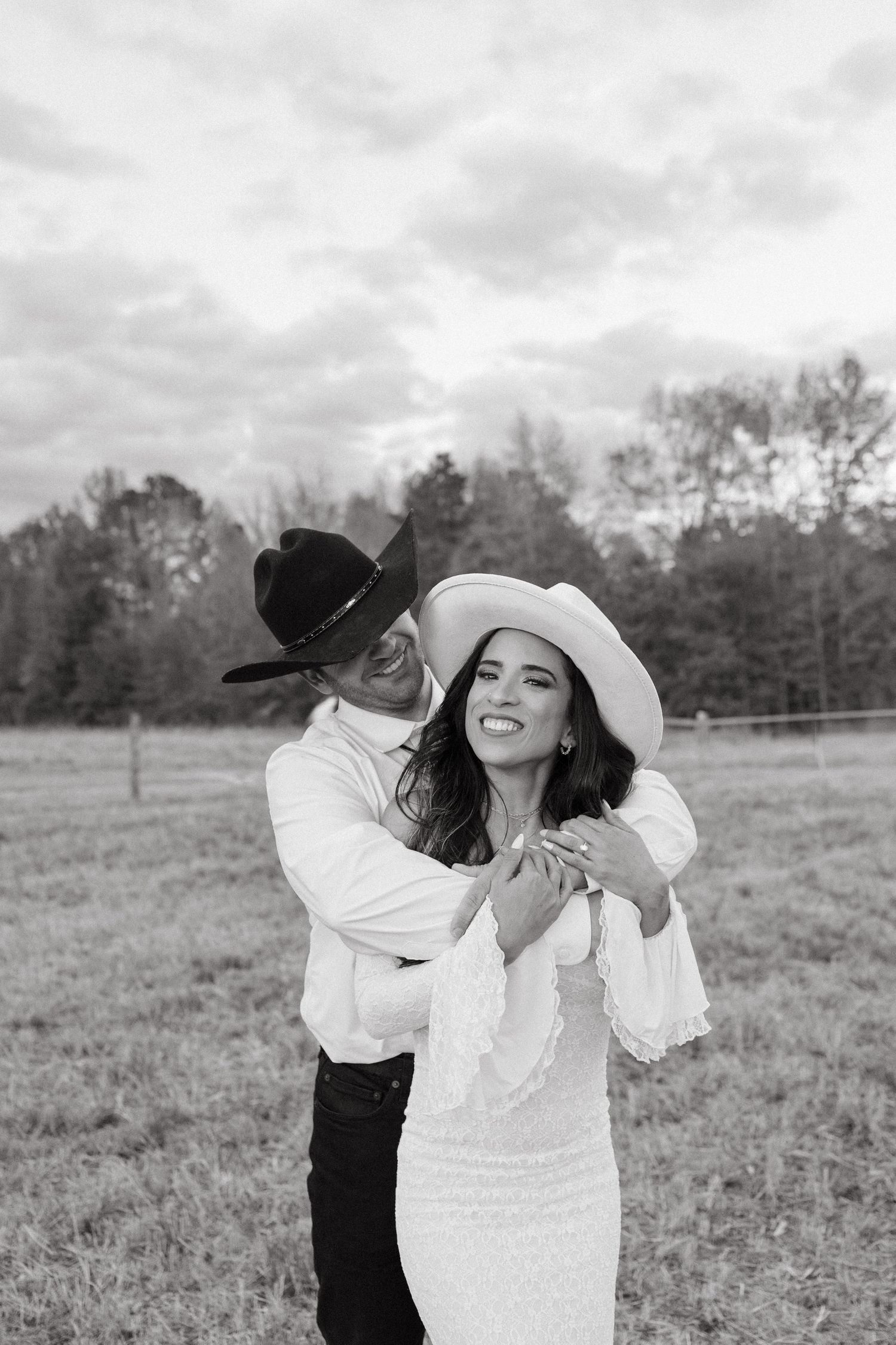 Black and white photo of groom embracing smiling bride wearing white cowboy hat in open field.