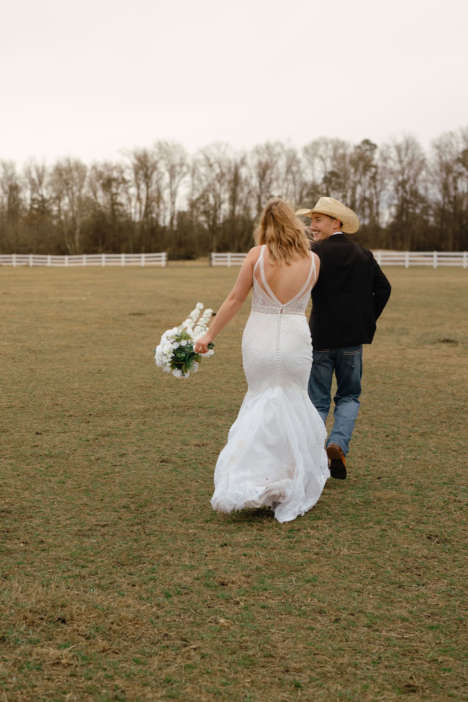 Bride in mermaid wedding gown and groom in cowboy hat walking through open field at farm venue.