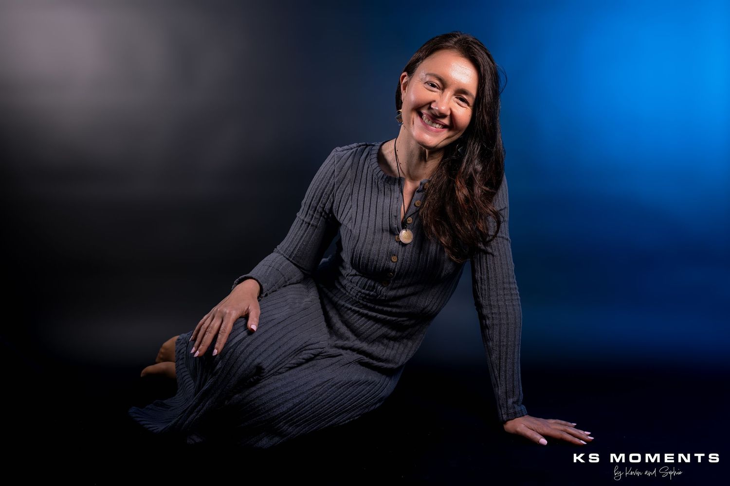 Photo d'une femme souriante assise par terre dans un studio photo avec un fond noir et des lumières de couleur bleue