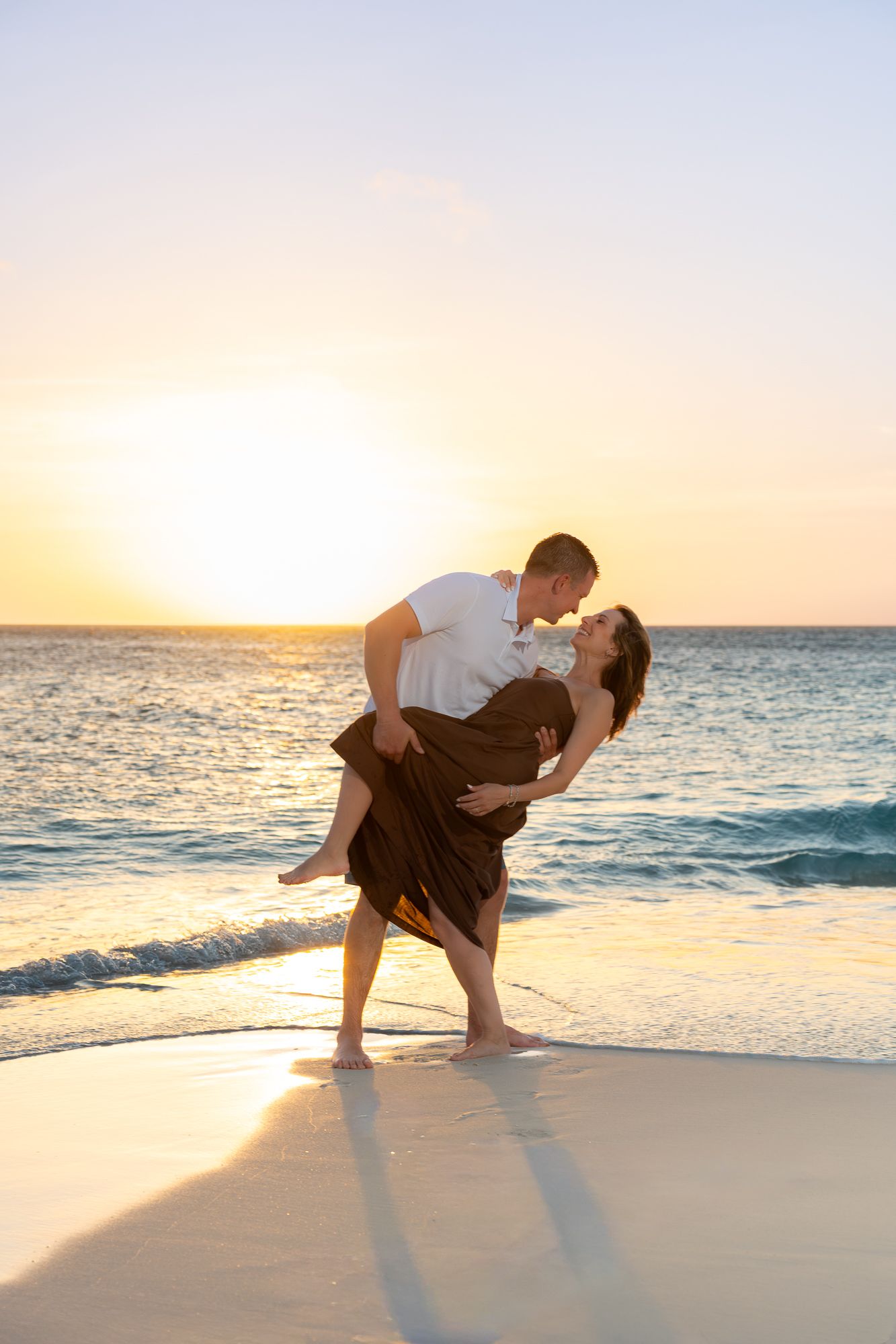 Man lifting woman in a romantic dip kiss on a sandy beach at vibrant golden sunset.